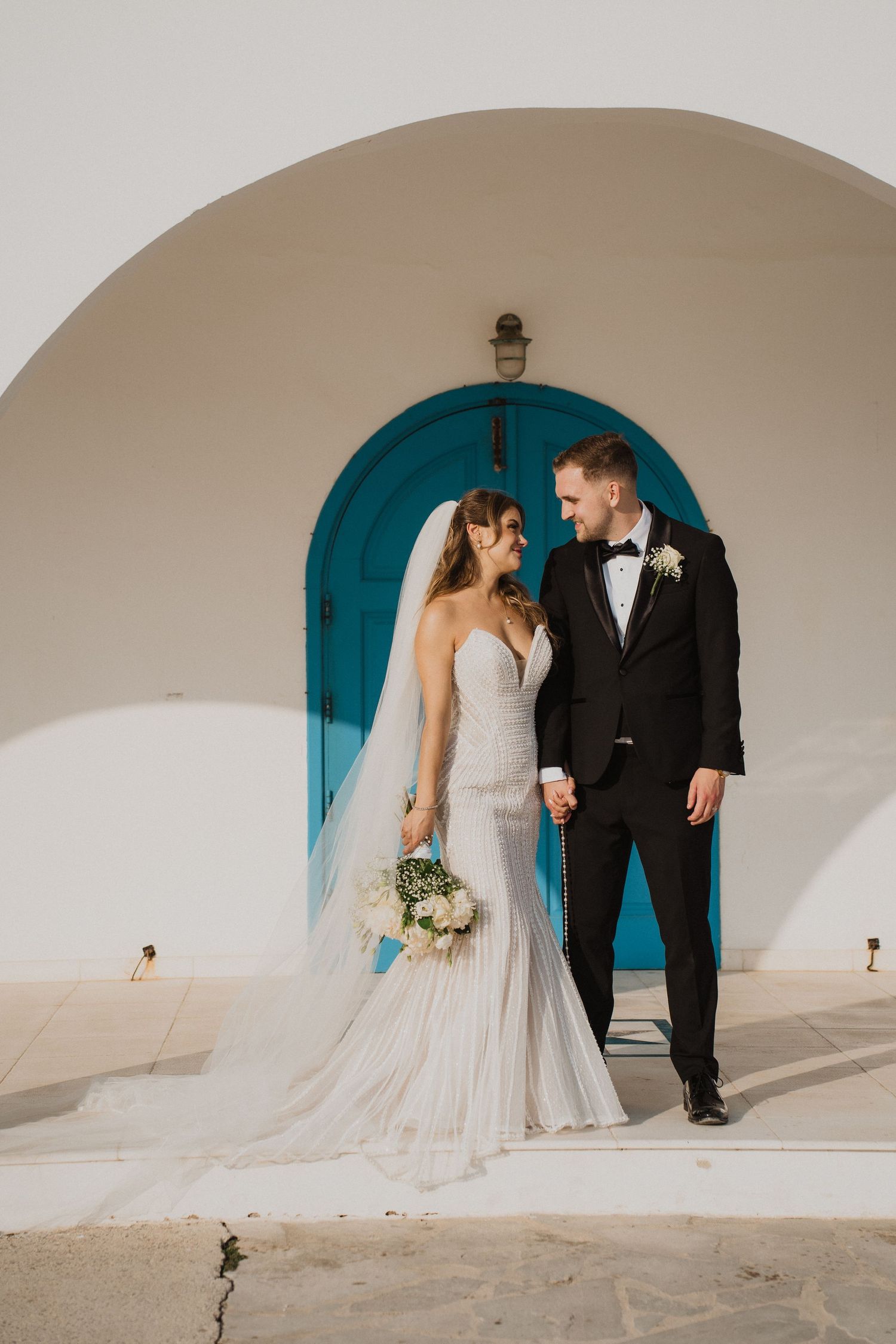 Bride and groom holding hands in front of a white arch with turquoise door at a Greek wedding venue.