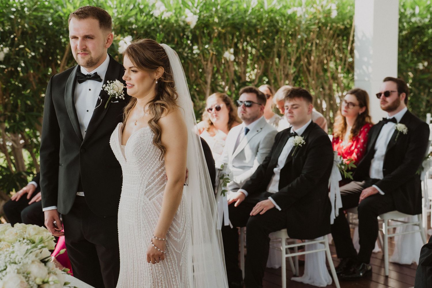 Bride and groom hold hands at outdoor garden wedding ceremony with seated guests watching.