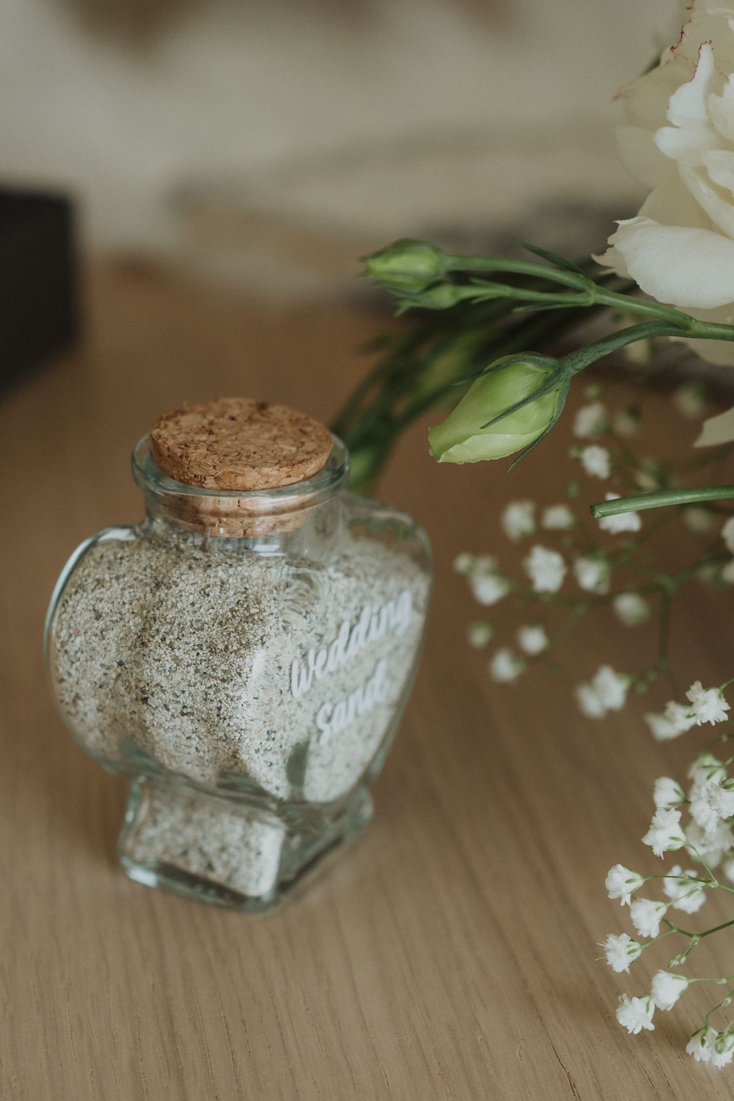Heart-shaped glass bottle filled with sand beside white wedding flowers.