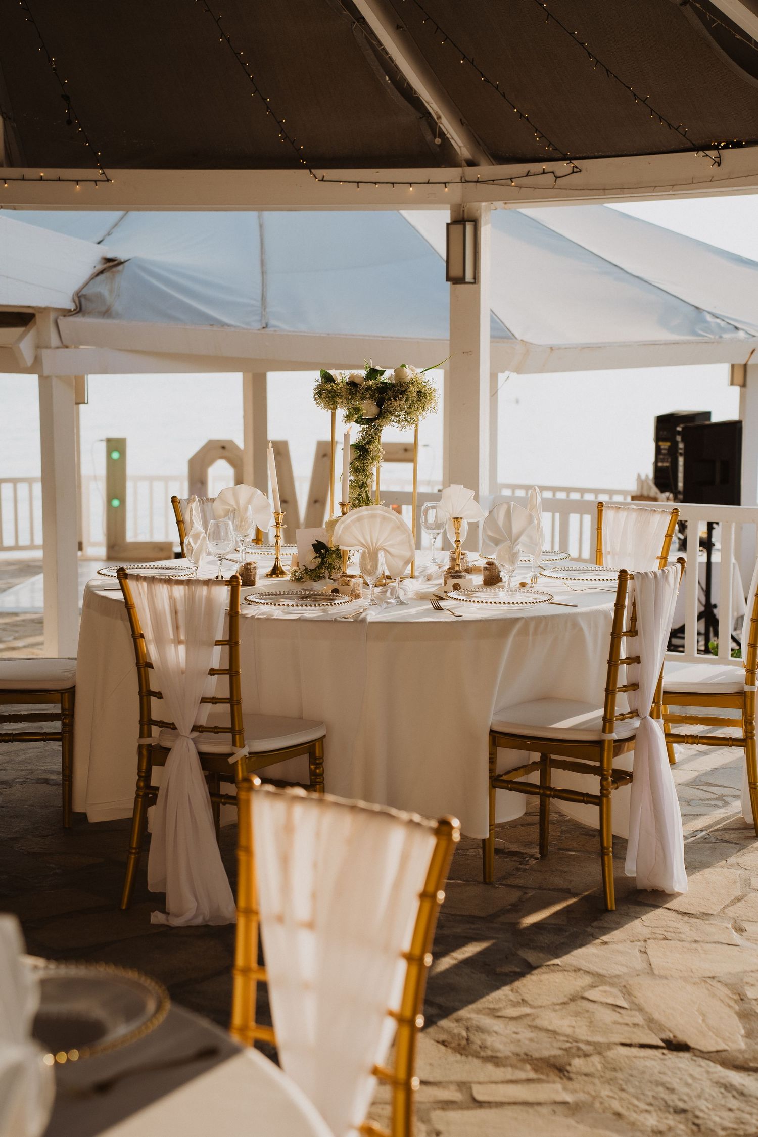 Seaside wedding reception gazebo with gold chiavari chairs, white linens, floral centerpieces and LOVE letters.