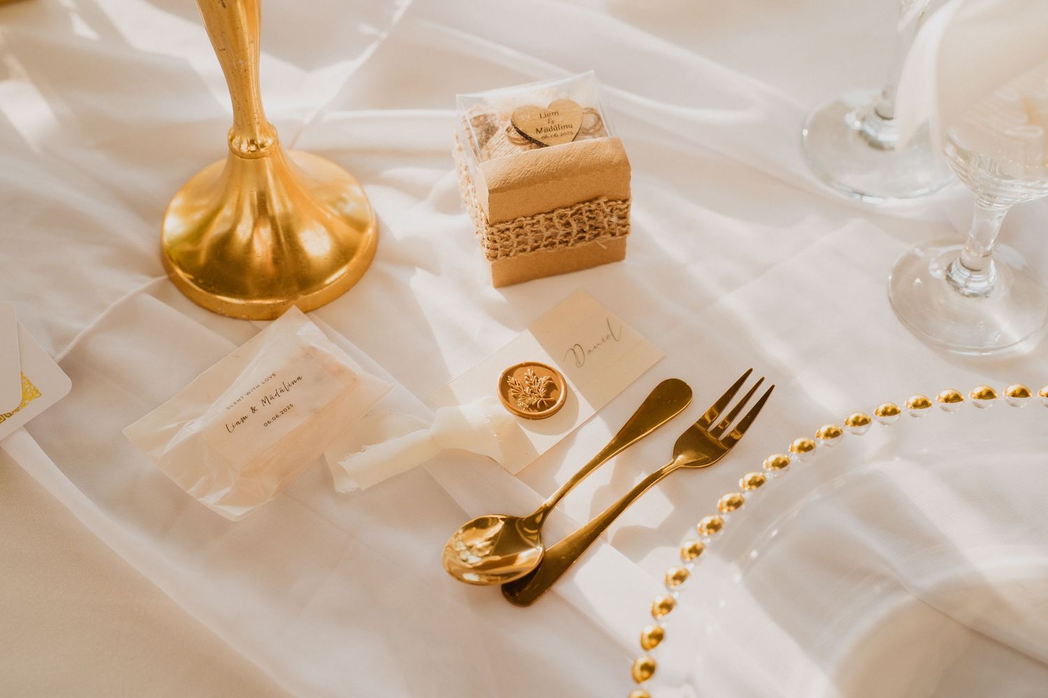 Elegant gold wedding place setting with wax seal, favor box, gold cutlery and beaded napkin on white linen.