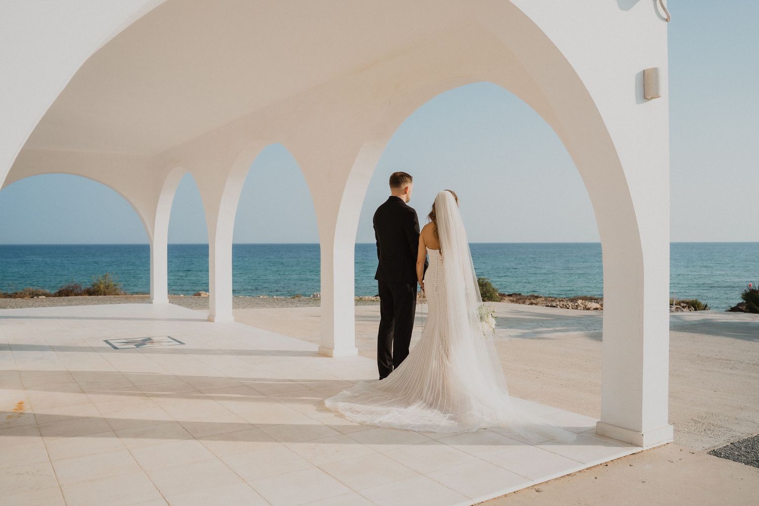 Bride and groom stand beneath white arched structure overlooking the blue ocean.
