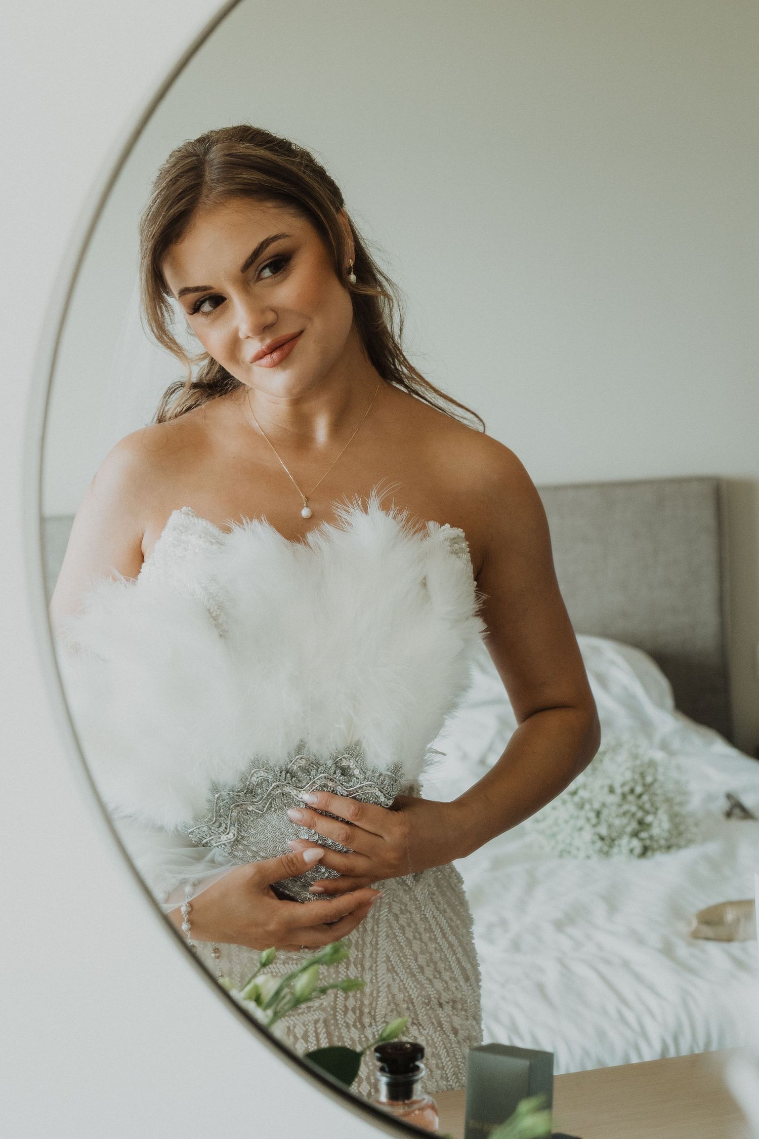 Bride in strapless beaded gown holding white feather bouquet reflected in round mirror.