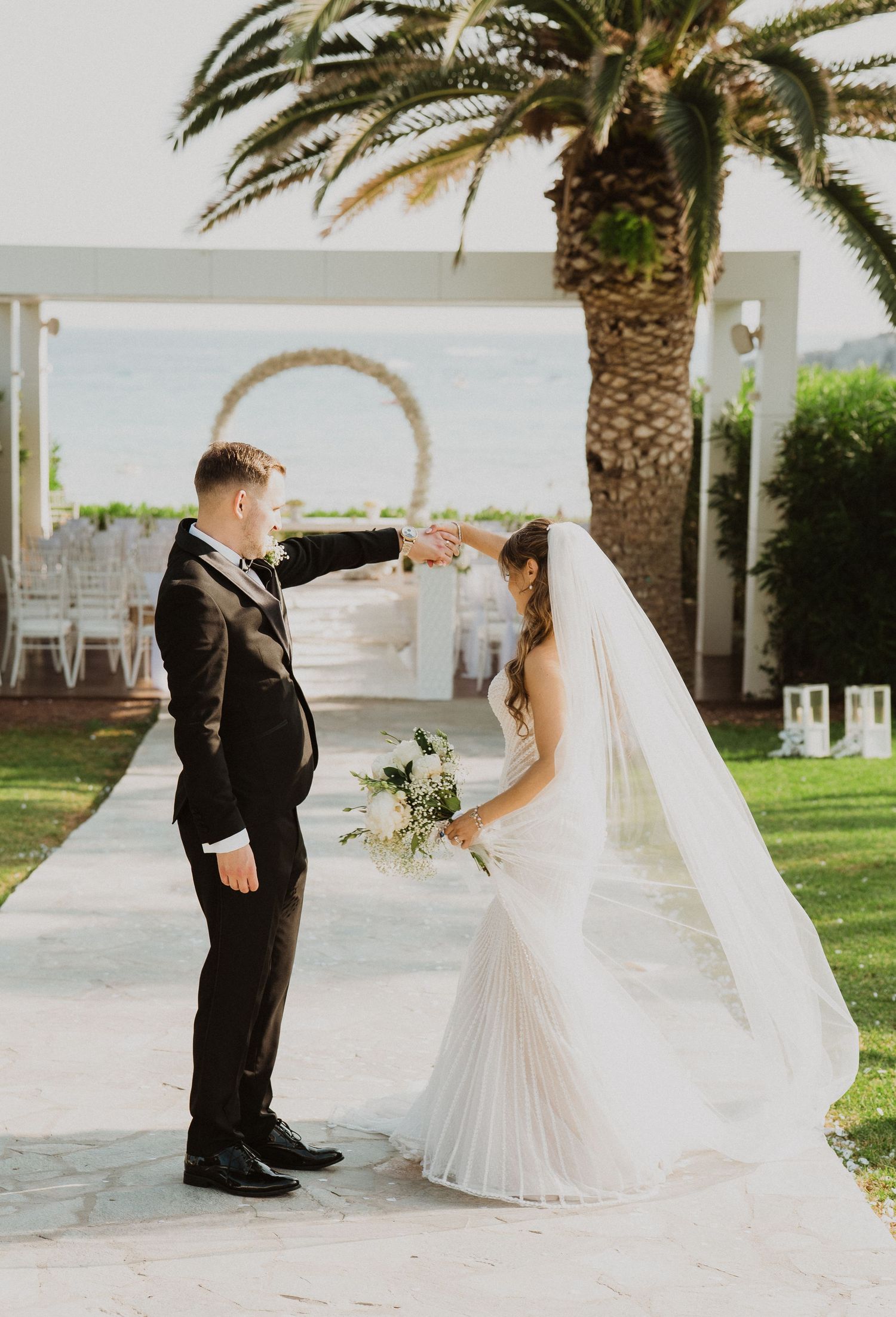 Groom twirling bride in white gown and veil on garden pathway with palm tree and ocean backdrop.