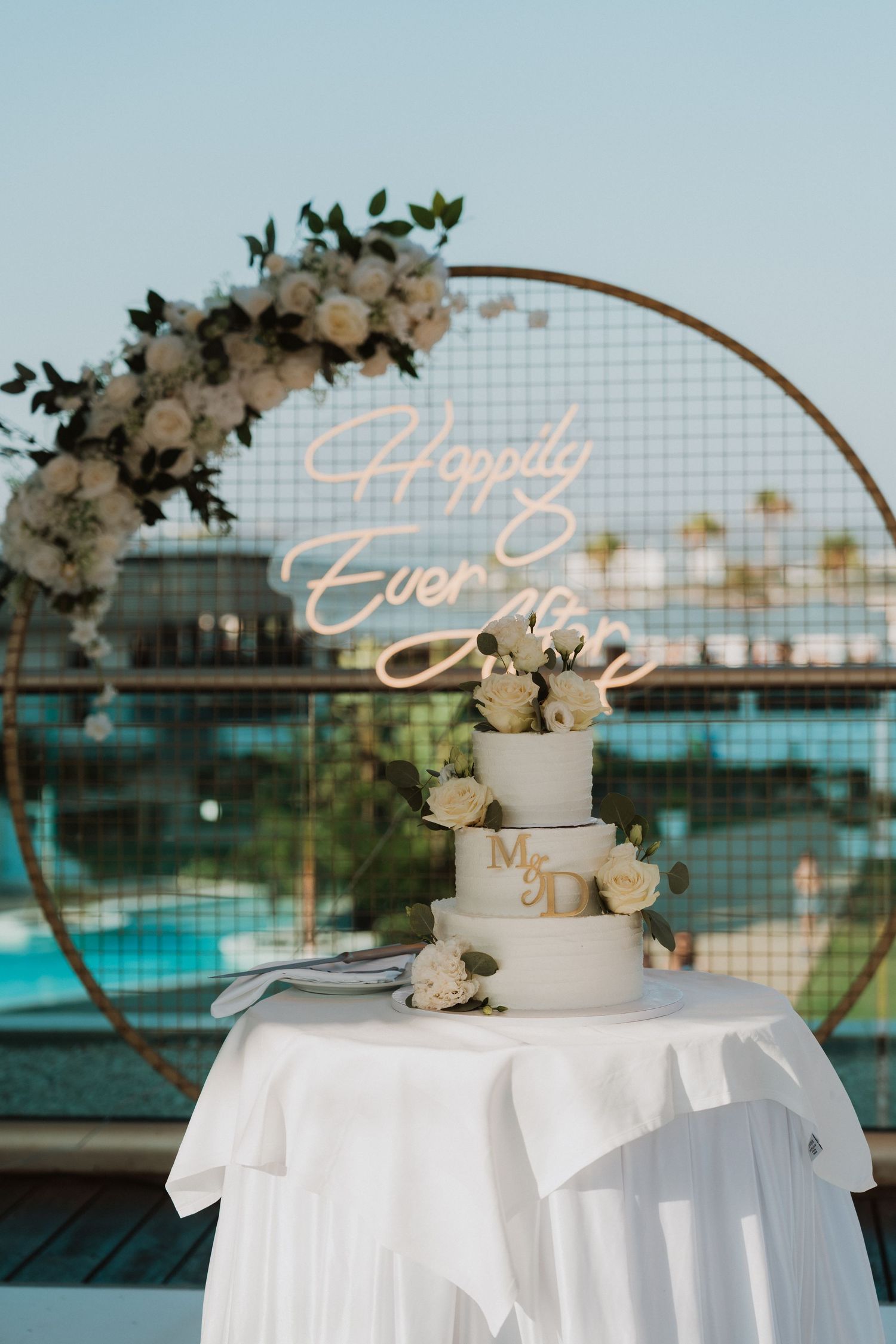 Elegant three-tier wedding cake with white roses and Mr & Mrs topper, set before a circular floral arch with Happily Ever After neon sign.