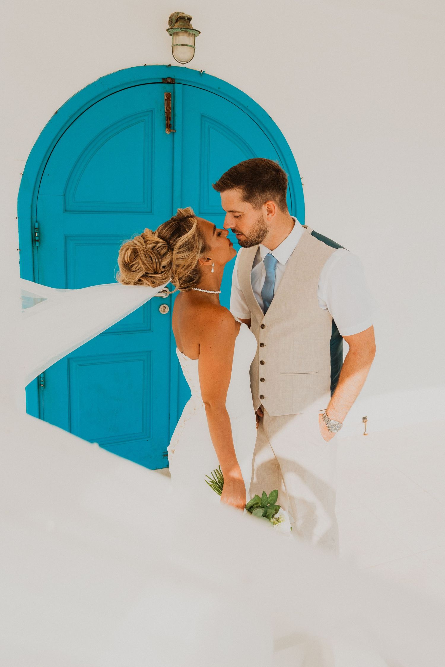 Bride and groom share a romantic moment in front of a vibrant turquoise arched door in Santorini.