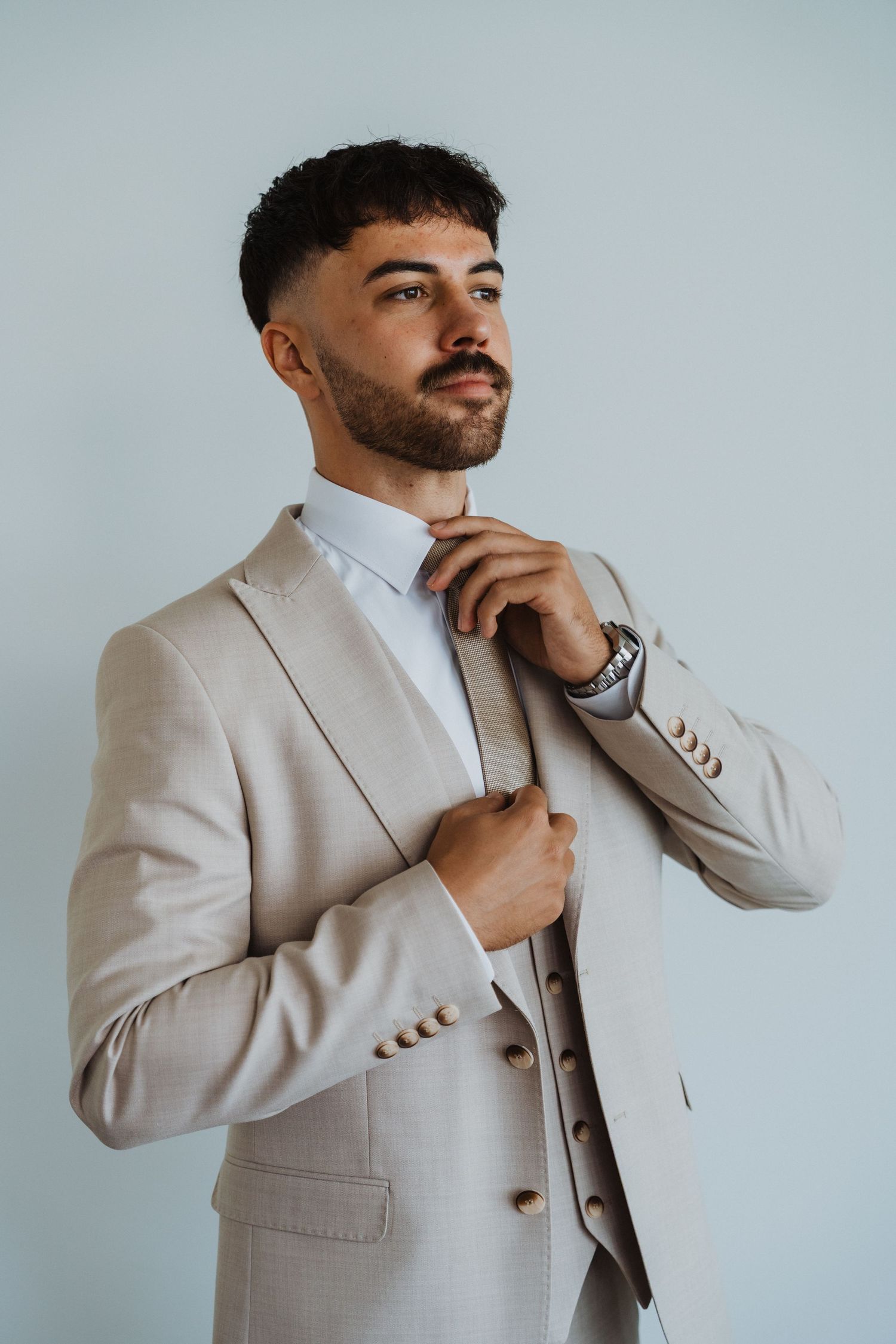 Man in beige wedding suit striking thoughtful poses during formal photoshoot.