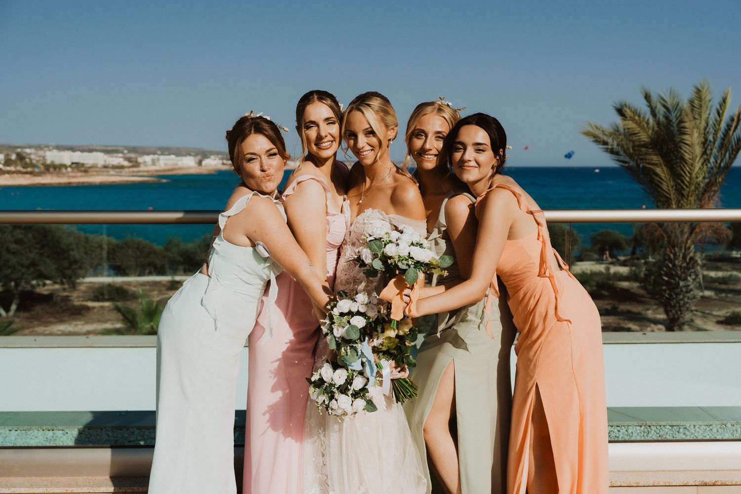 Bridesmaids in soft-colored dresses embrace on a coastal balcony.
