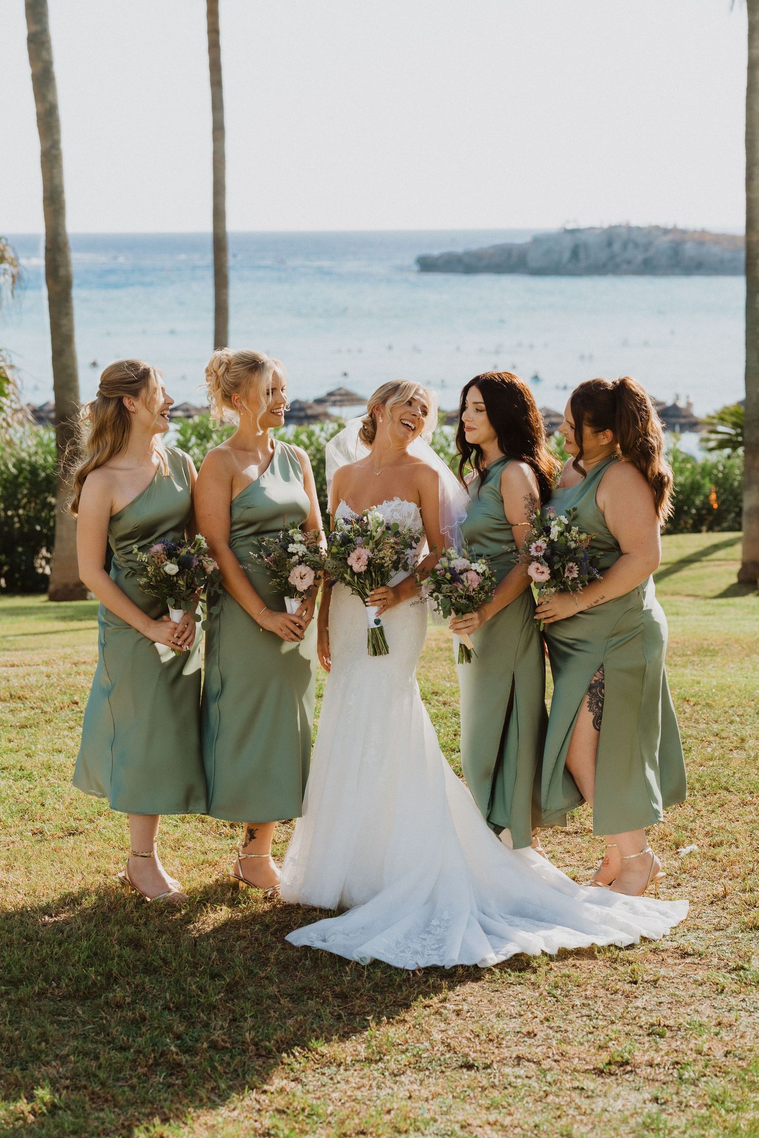 Bridesmaids in sage dresses gather around bride for photos with ocean view and palm trees.