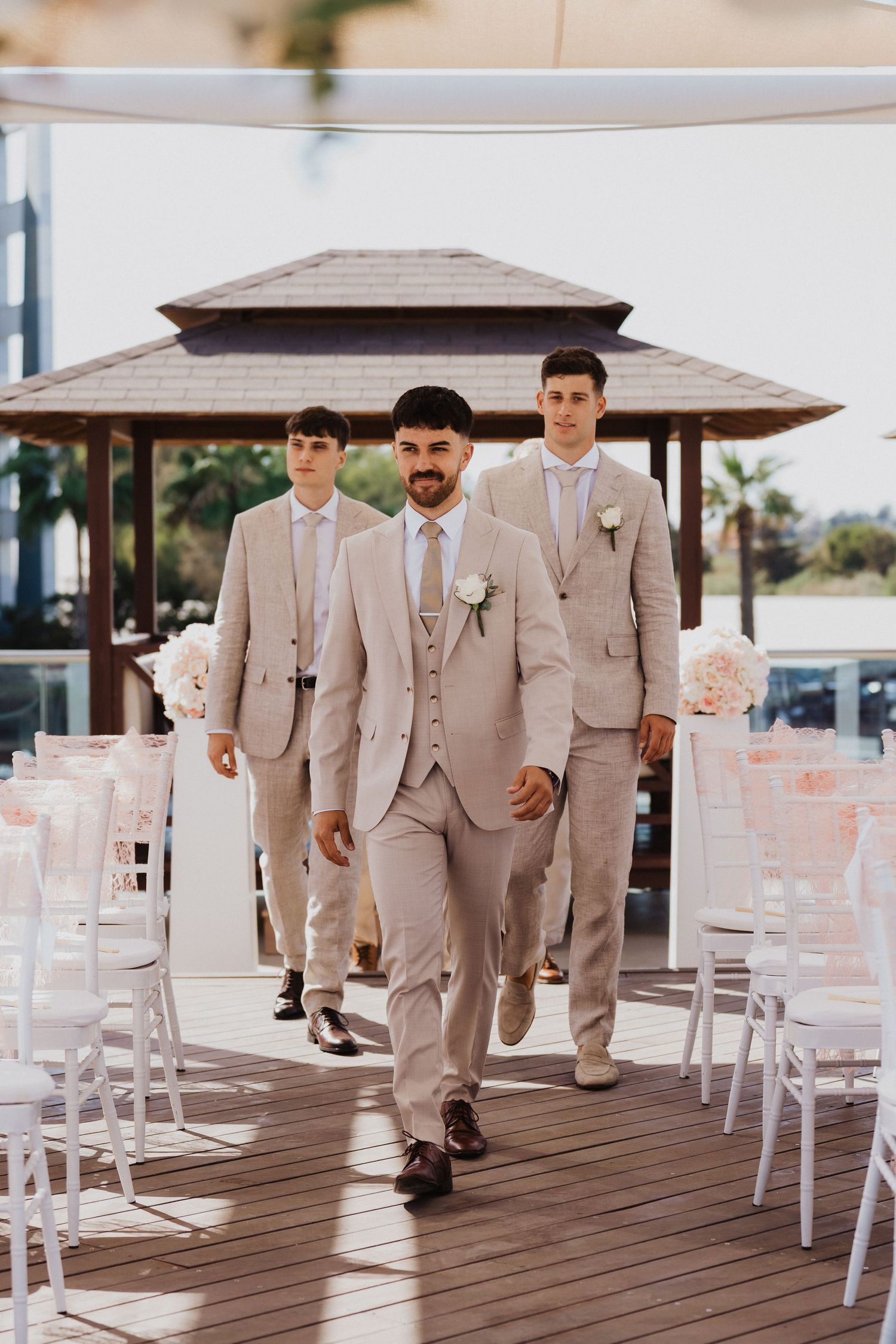 Wedding party members in light suits walk through an outdoor venue with white chairs.