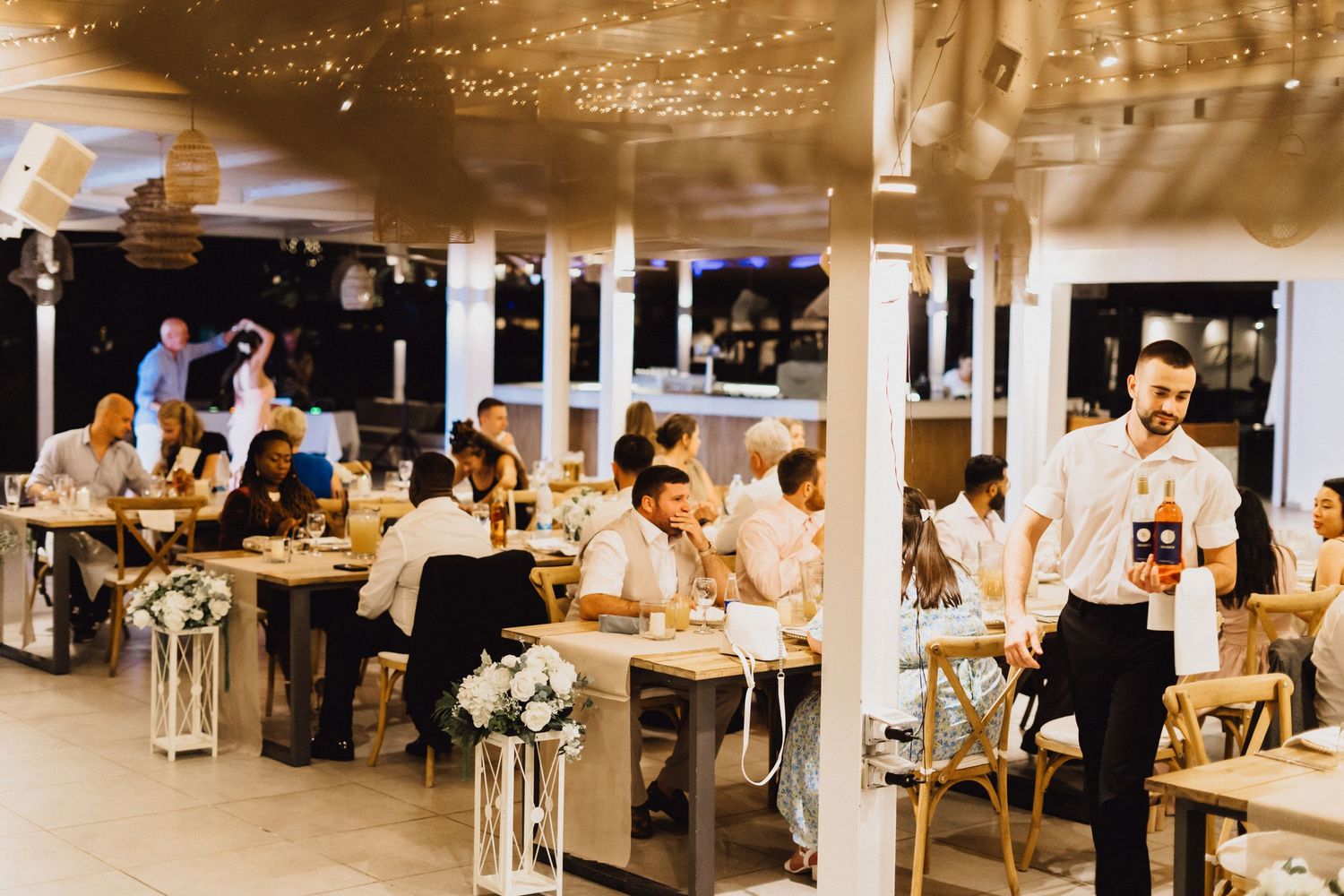 Wedding reception venue with guests seated at tables under string lights and white ceiling beams.