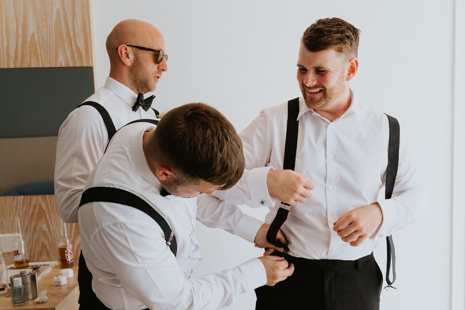 Groomsmen in white shirts and suspenders share a lighthearted moment while getting ready.