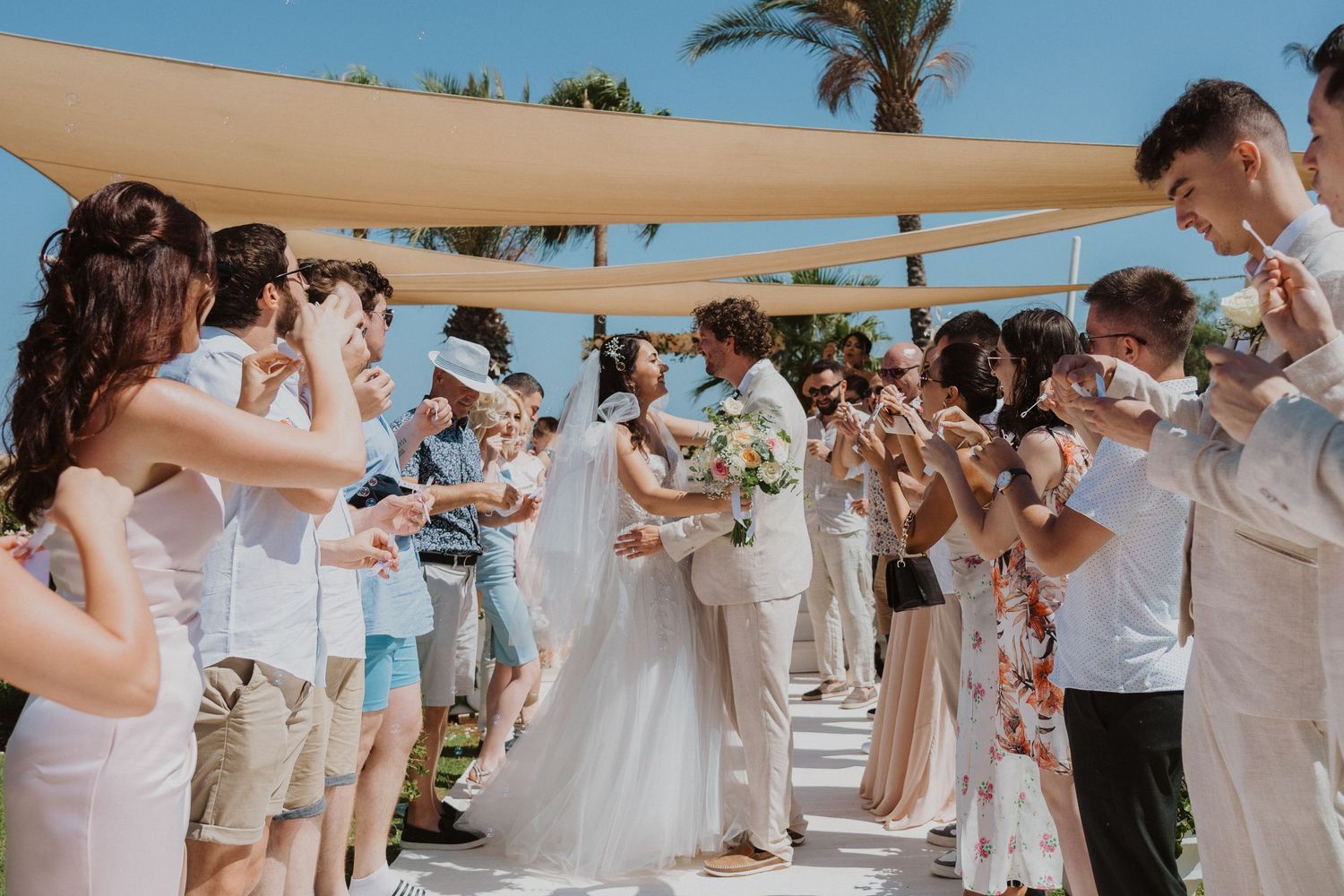 Wedding guests create celebratory bubble send-off at beach ceremony under shade sails.