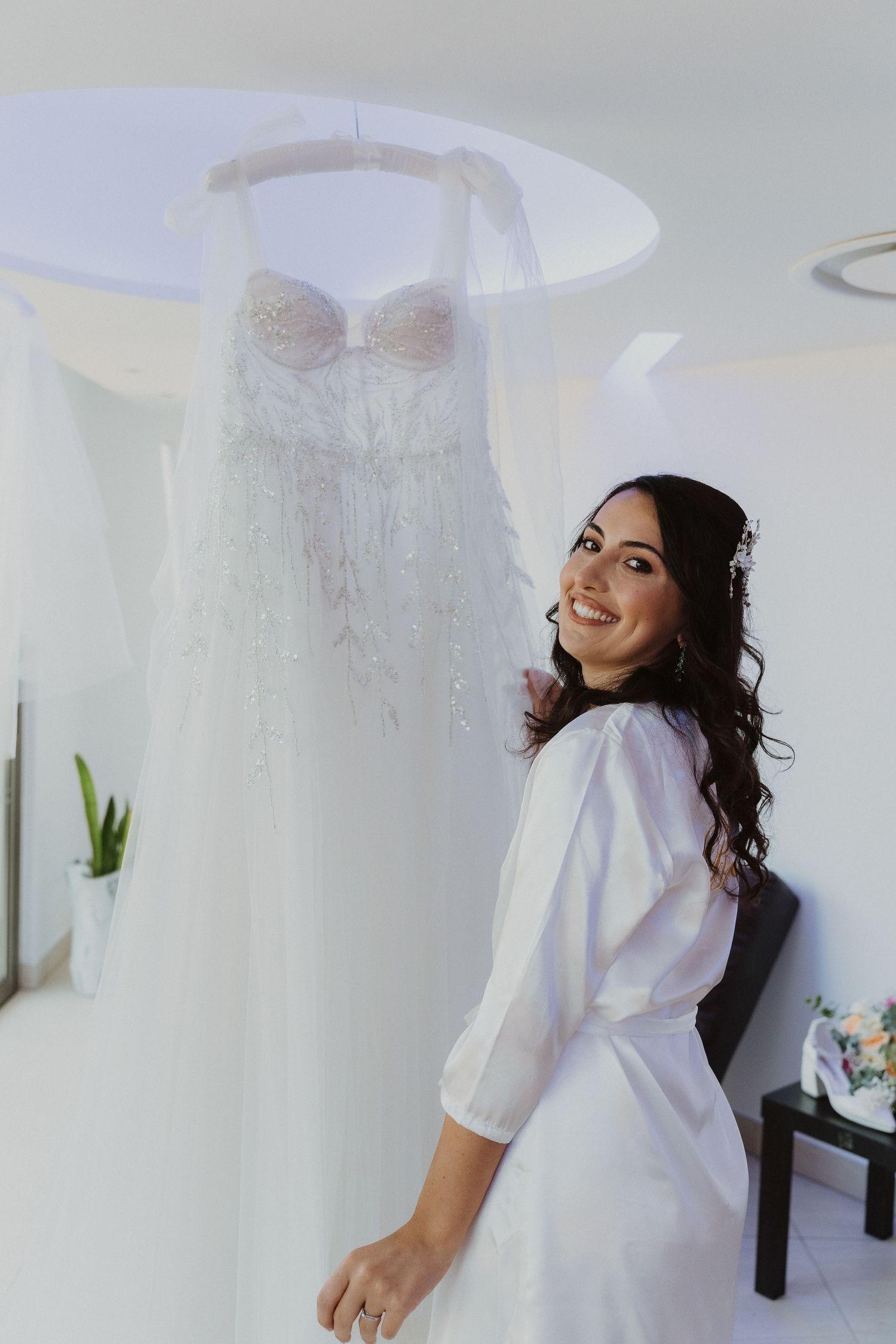 Bride in white robe smiles while looking at her wedding dress hanging in a bright dressing room.