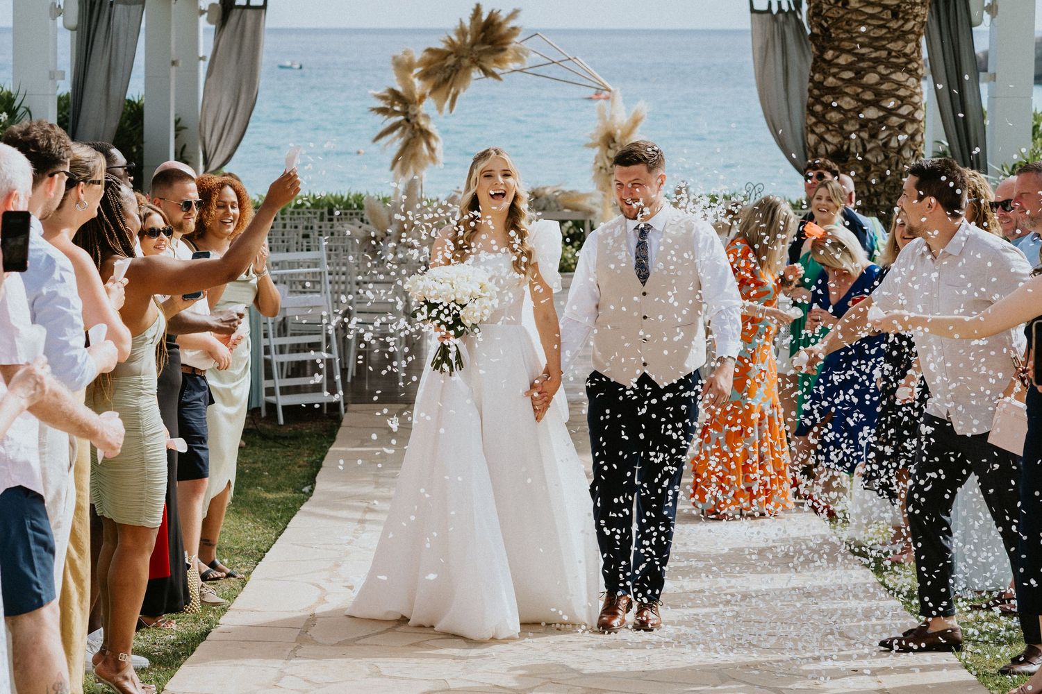 Joyful couple walks down outdoor aisle at beach wedding while guests throw confetti in celebration.