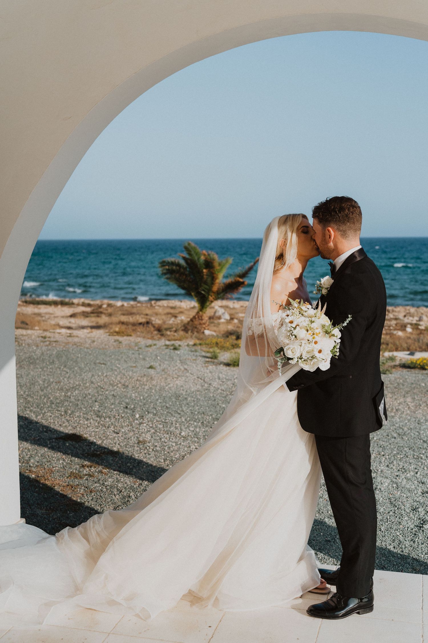 A tender seaside wedding portrait under white arches with ocean views and palm trees.