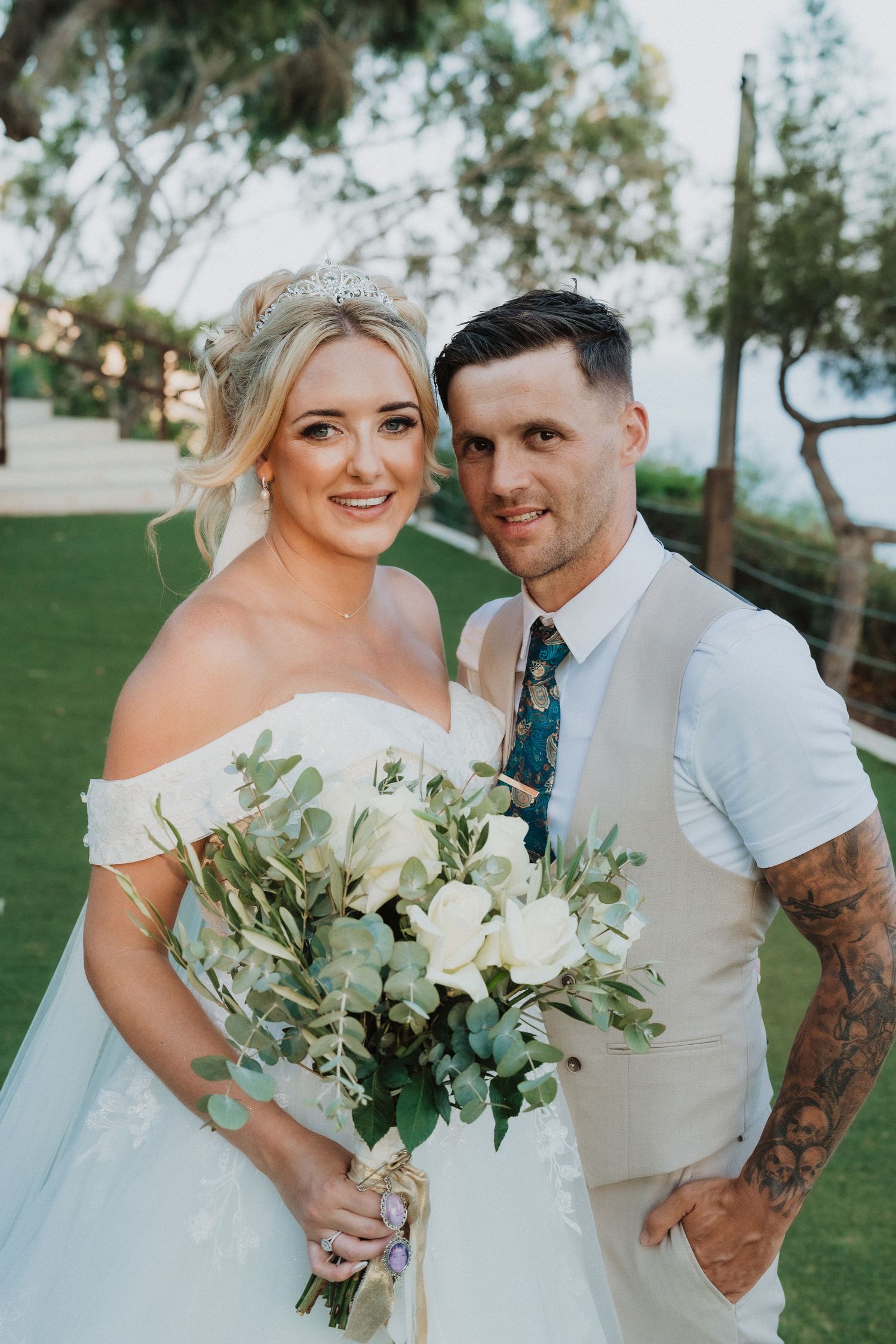 A romantic outdoor wedding photo featuring a couple with a beautiful white floral bouquet on a grassy lawn.