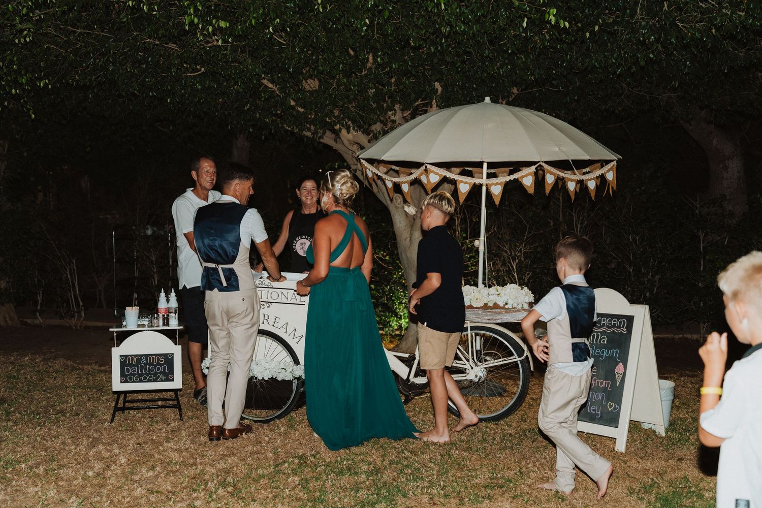 Evening outdoor gathering with guests near a white umbrella cart and decorative lights.