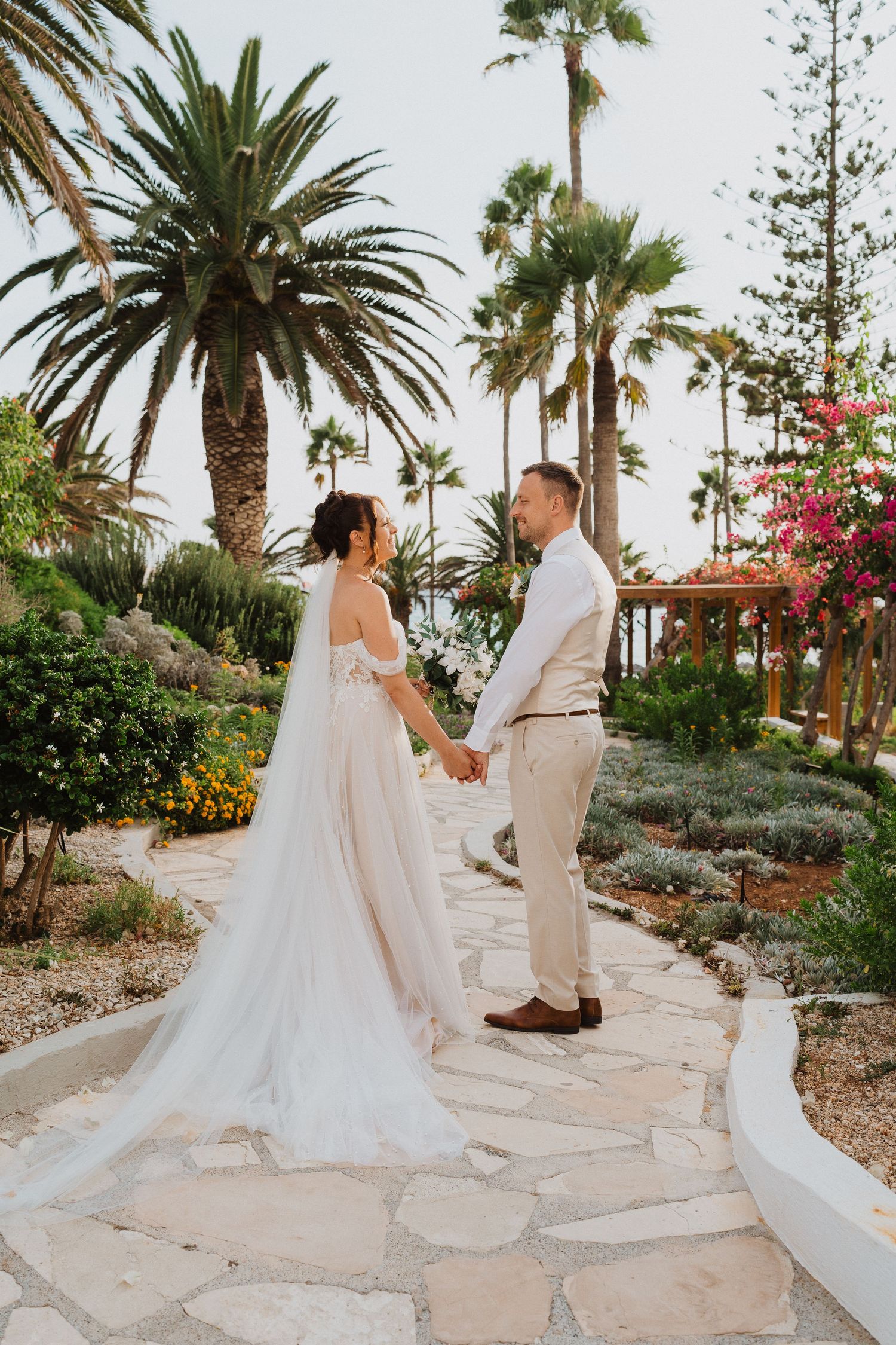 A couple shares a romantic moment on a stone path surrounded by palm trees and tropical landscaping at their wedding.