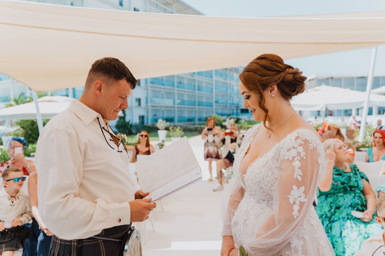 A romantic outdoor wedding scene with a couple exchanging vows by the ocean under a white tent.