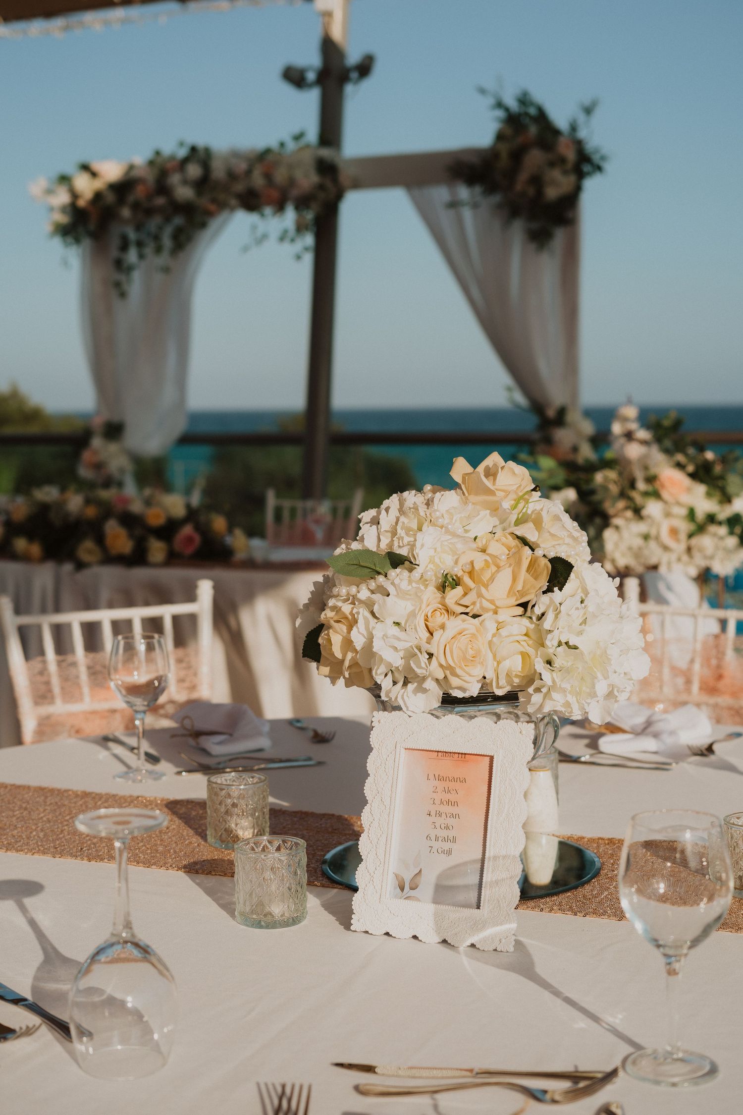 Elegant wedding table setting with floral centerpiece and draped ceremony arch overlooking ocean.