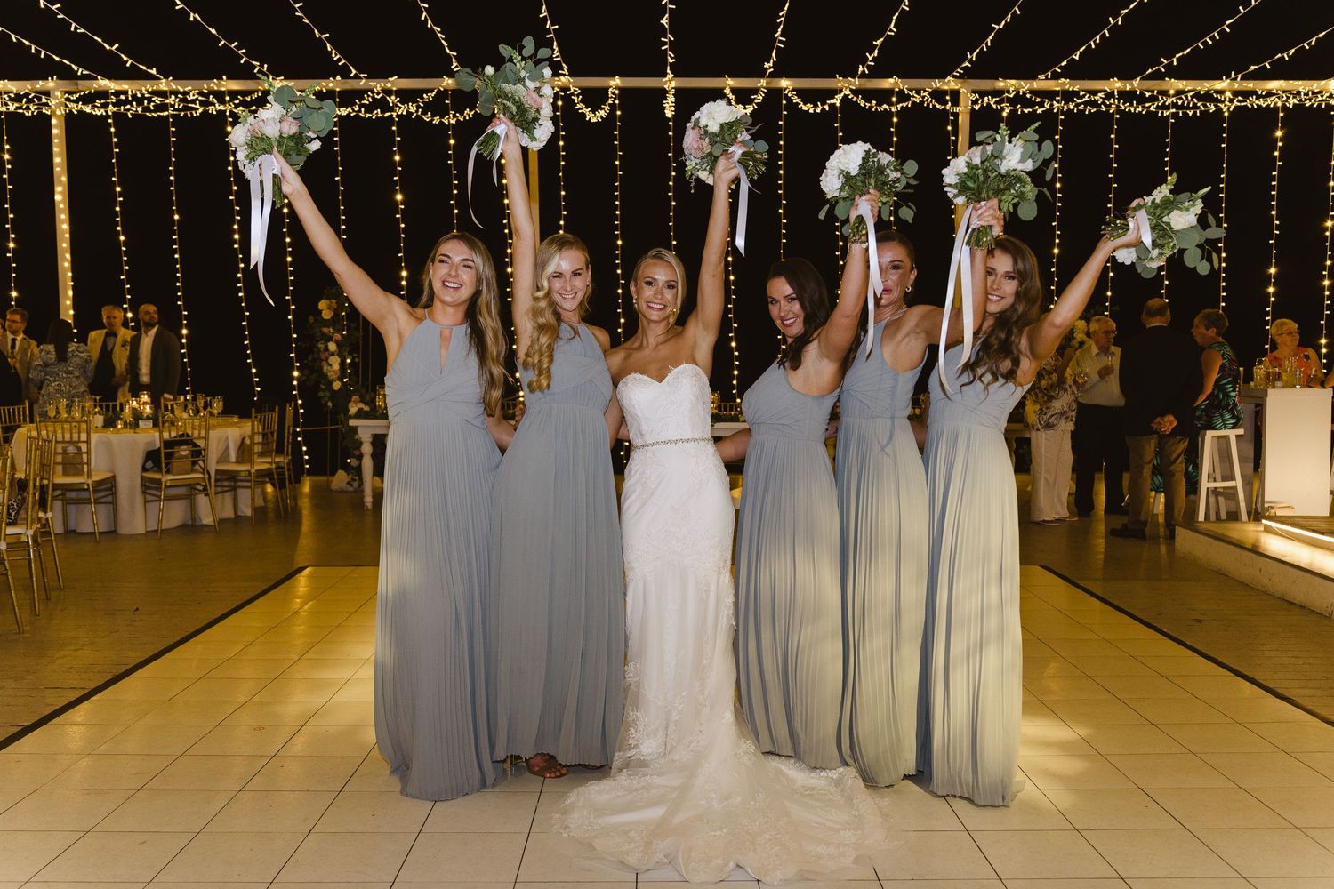 Bridesmaids in grey dresses raise bouquets with bride in white gown under string lights.