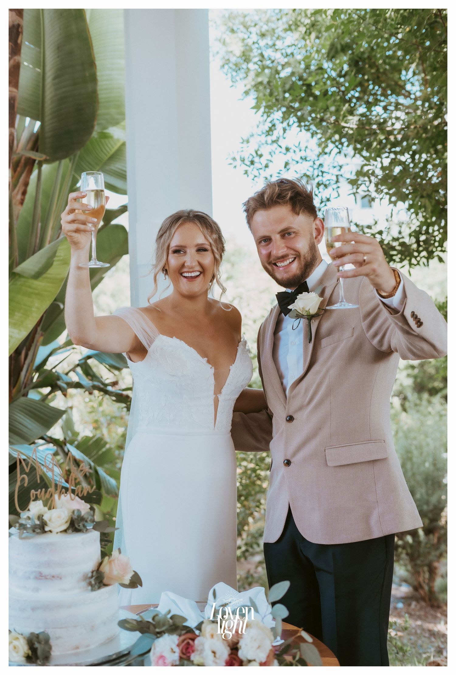 Couple raises champagne glasses in celebration next to their wedding cake outdoors.