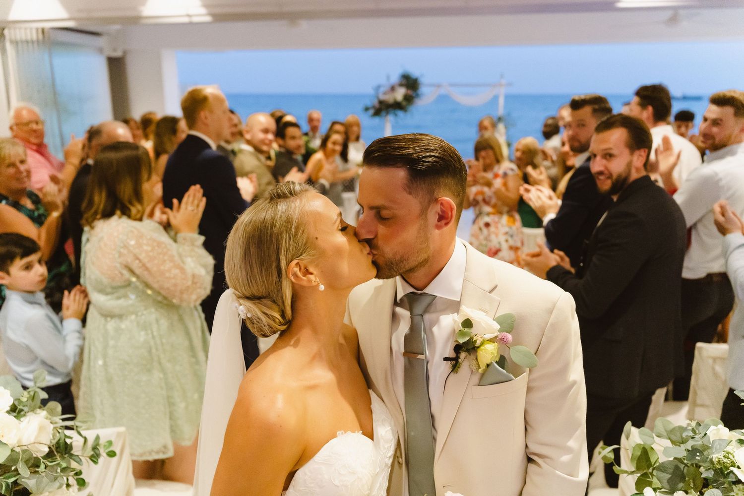 Newlyweds share a romantic kiss during their wedding reception surrounded by cheering guests.