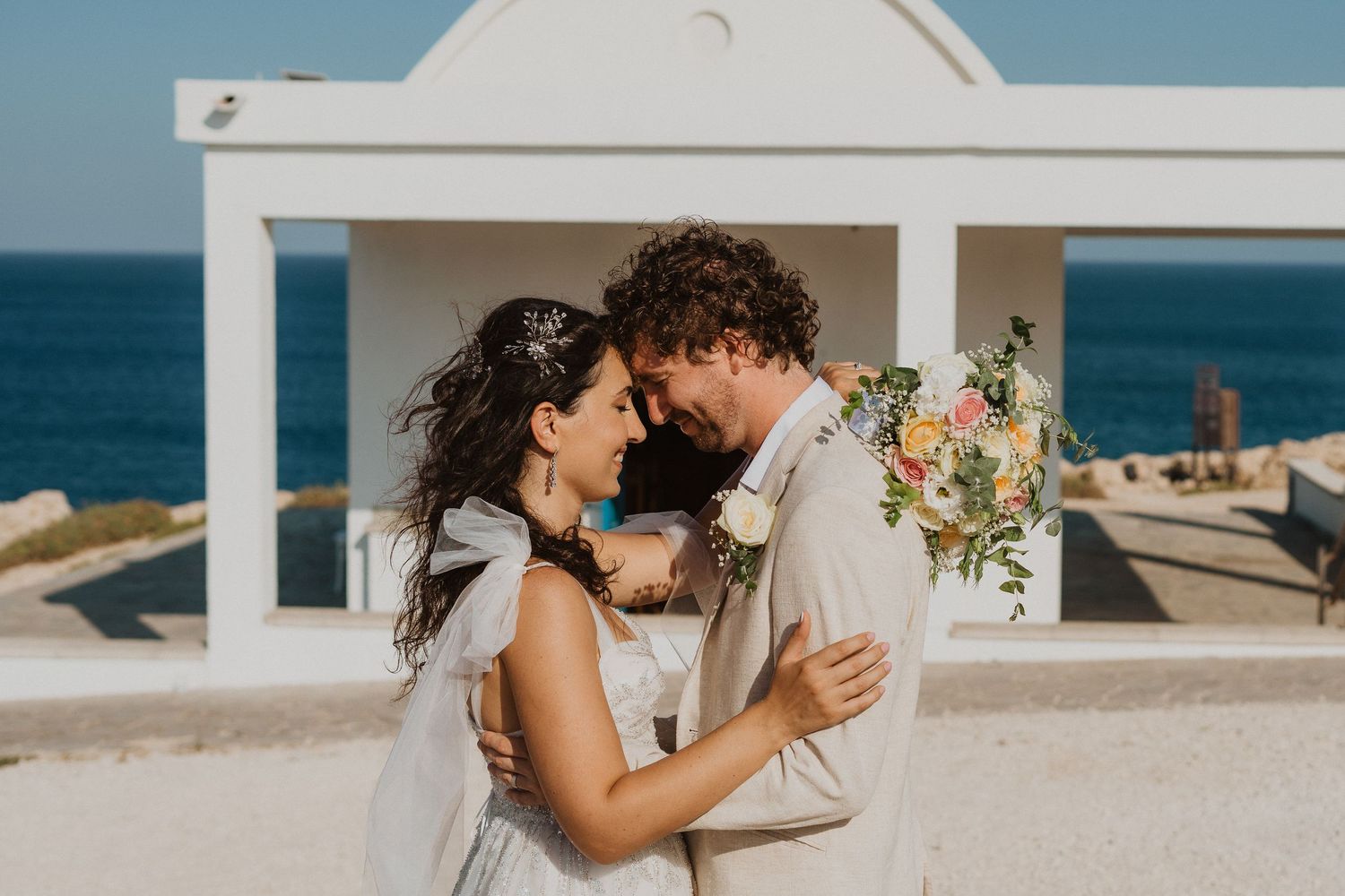 A loving embrace between newlyweds outside a white coastal chapel with ocean views.