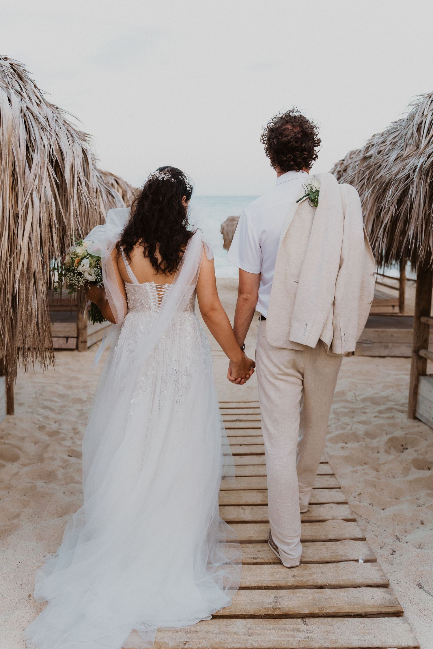 Newlyweds walk hand in hand on a wooden beach boardwalk surrounded by palm frond umbrellas.