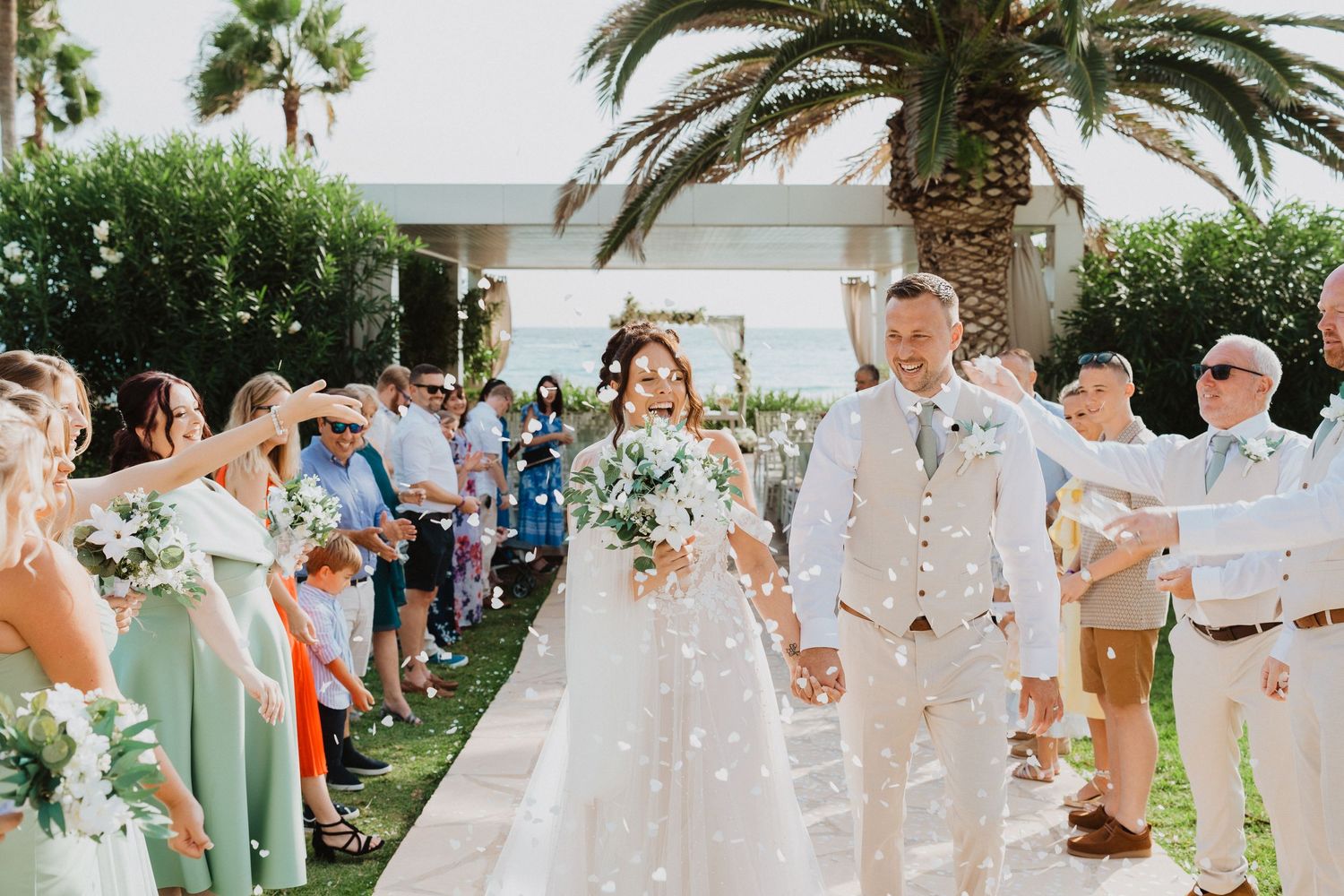 Joyful wedding couple walks through a palm tree lined path as guests celebrate and toss confetti at an outdoor ceremony.