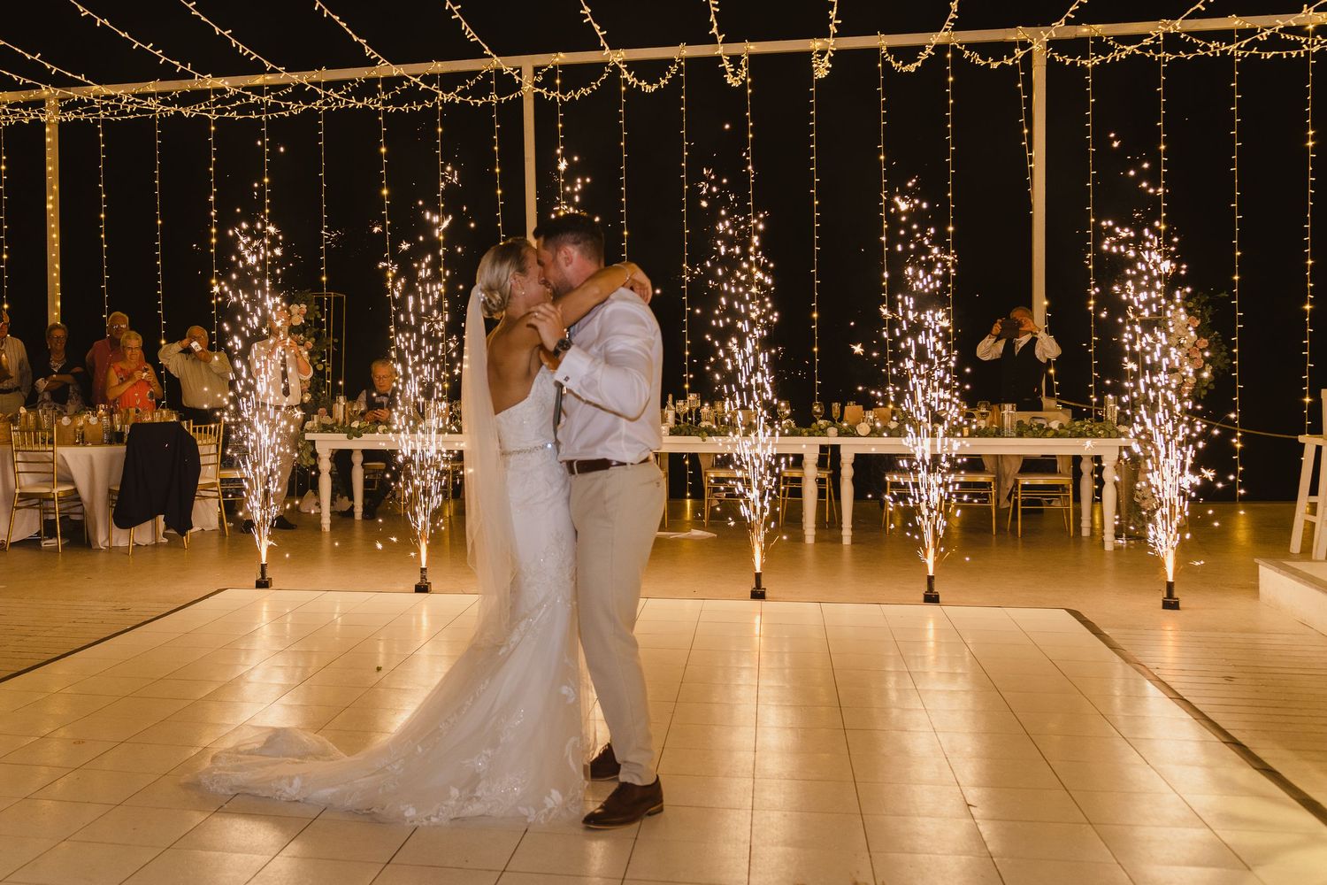 Couple shares romantic first dance surrounded by sparkling indoor fireworks and string lights at wedding reception.