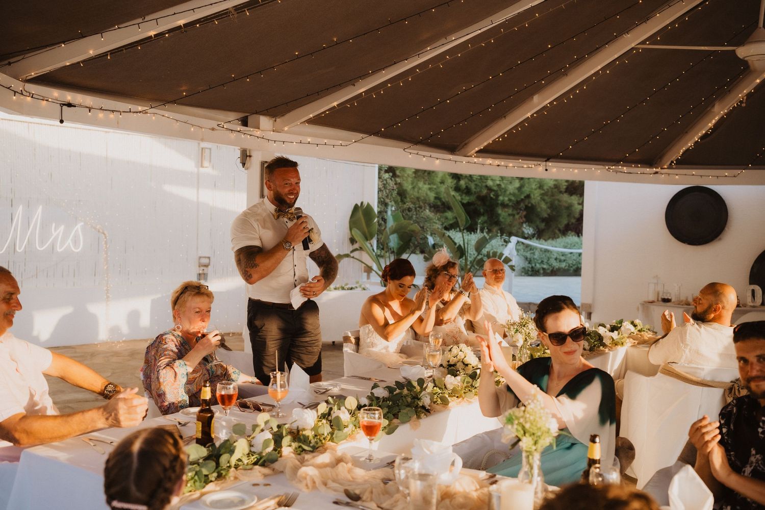 Wedding reception guests gathered around decorated dinner table during celebratory speeches.