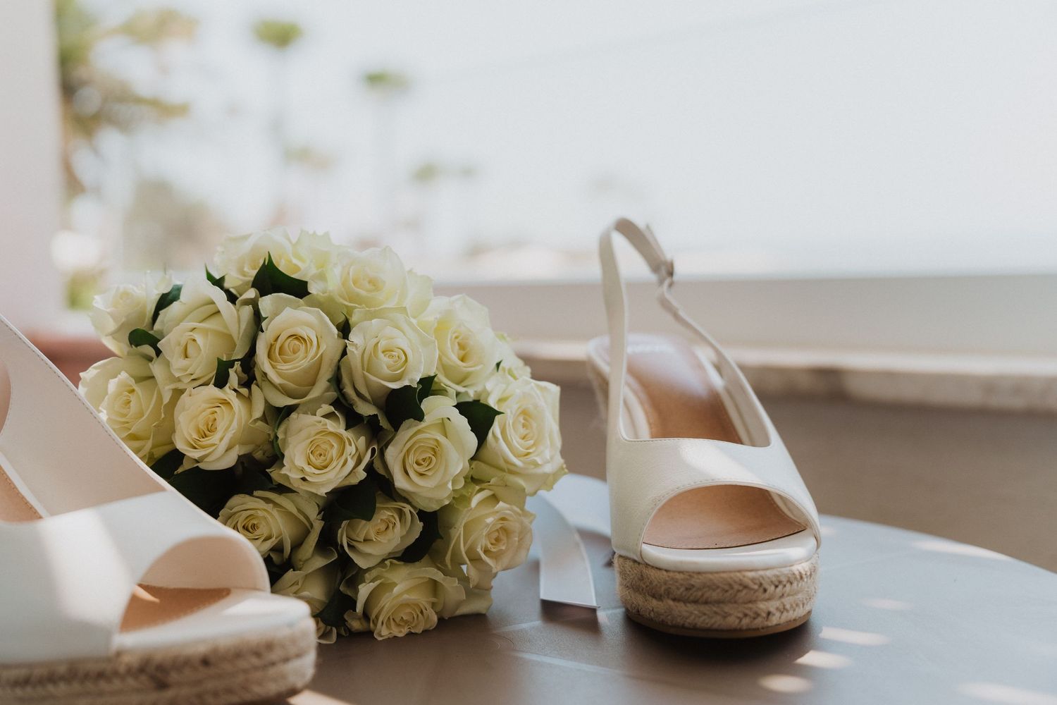 White bridal shoes and cream rose bouquet arranged on coastal balcony.