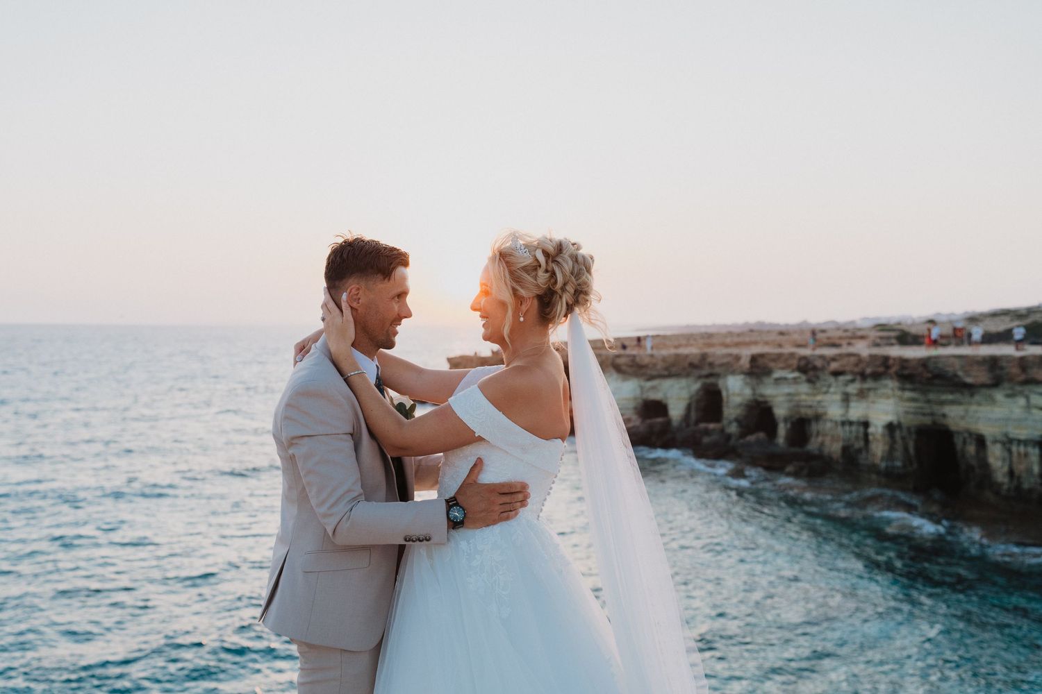 A romantic wedding couple embraces on a seaside cliff at sunset, with the ocean and rocky coastline in the background.