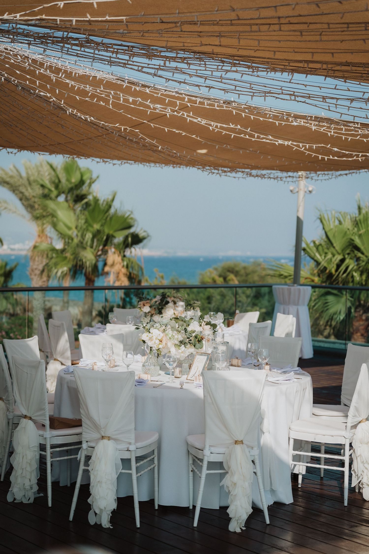 Elegant white wedding reception tables with draped chairs under strings lights by the sea.
