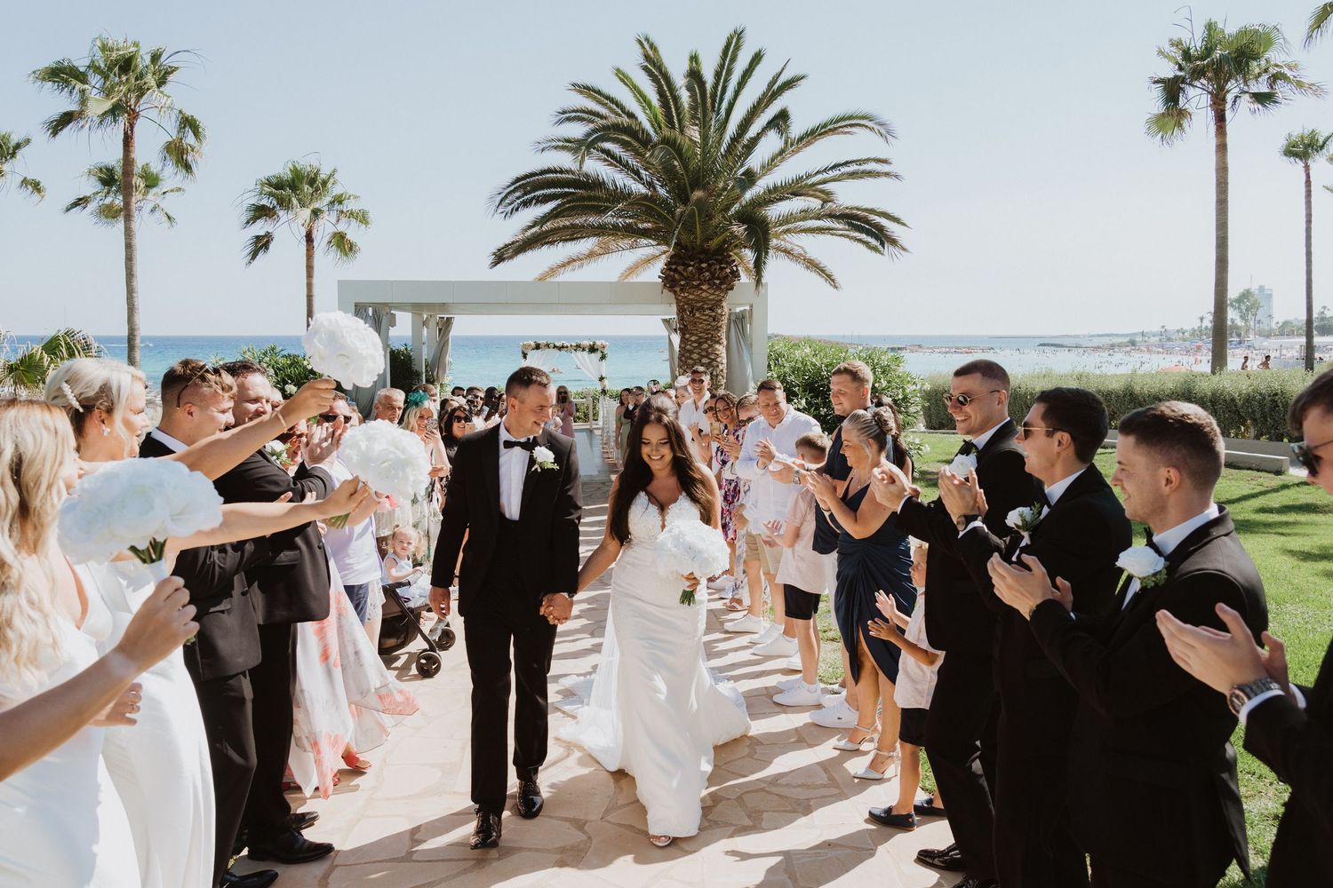 Wedding guests lined up along a palm-lined pathway by the ocean for a celebration.