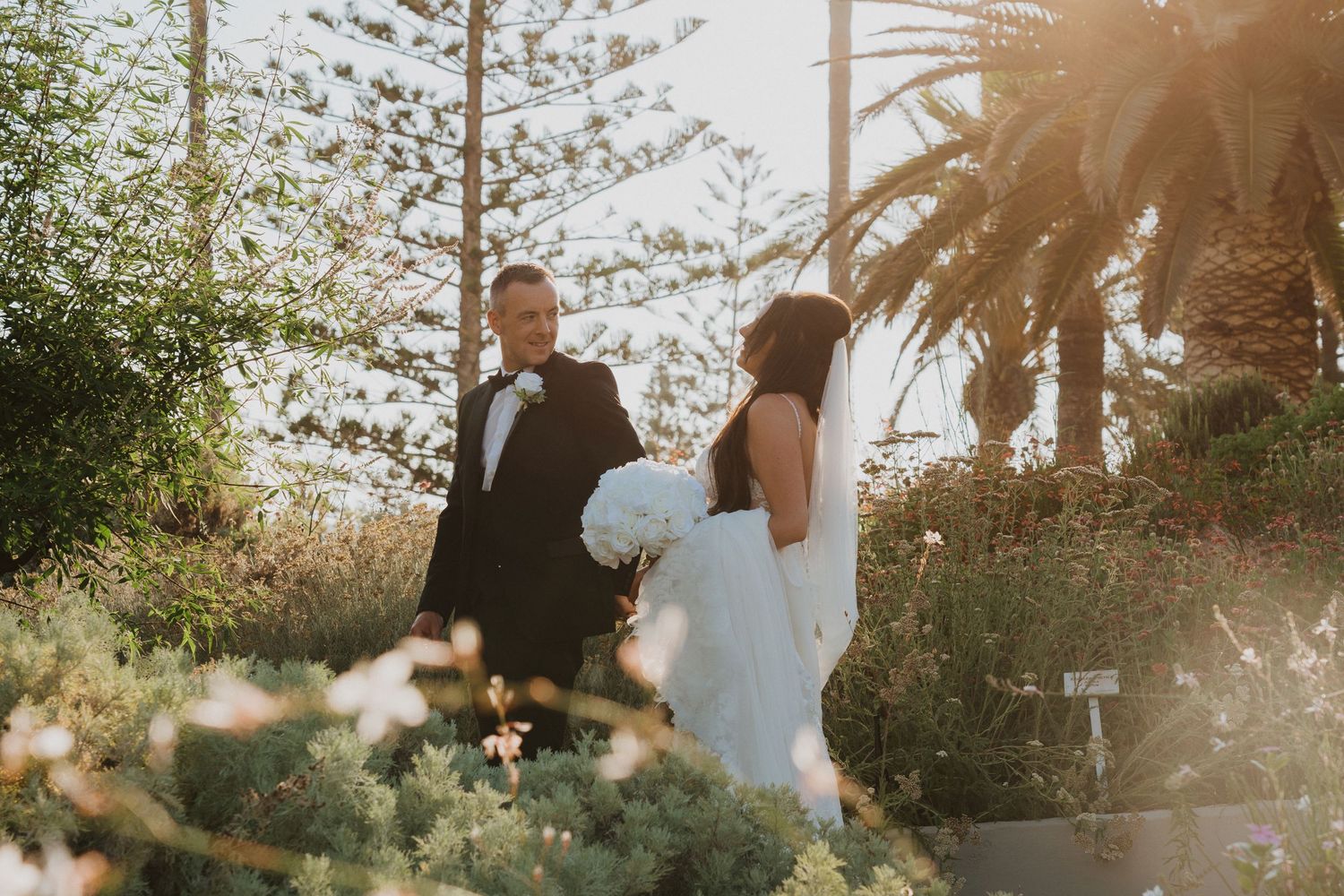A couple in wedding attire strolls through a garden path with palm trees in the background.