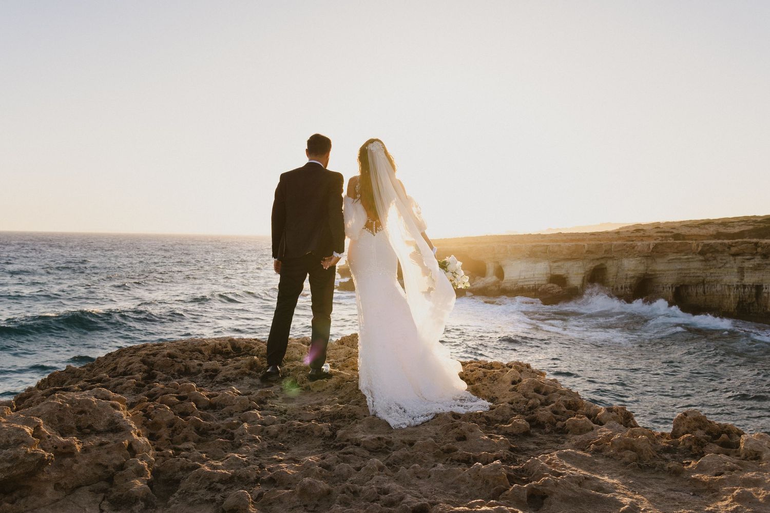 Couple stands on rocky coastal cliff overlooking Mediterranean Sea at sunset.