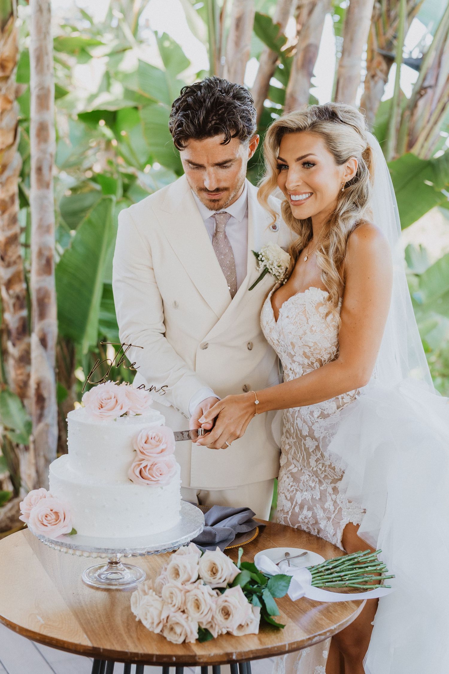 A newlywed couple cuts their elegant white wedding cake decorated with pink roses on a wooden table in a tropical setting.
