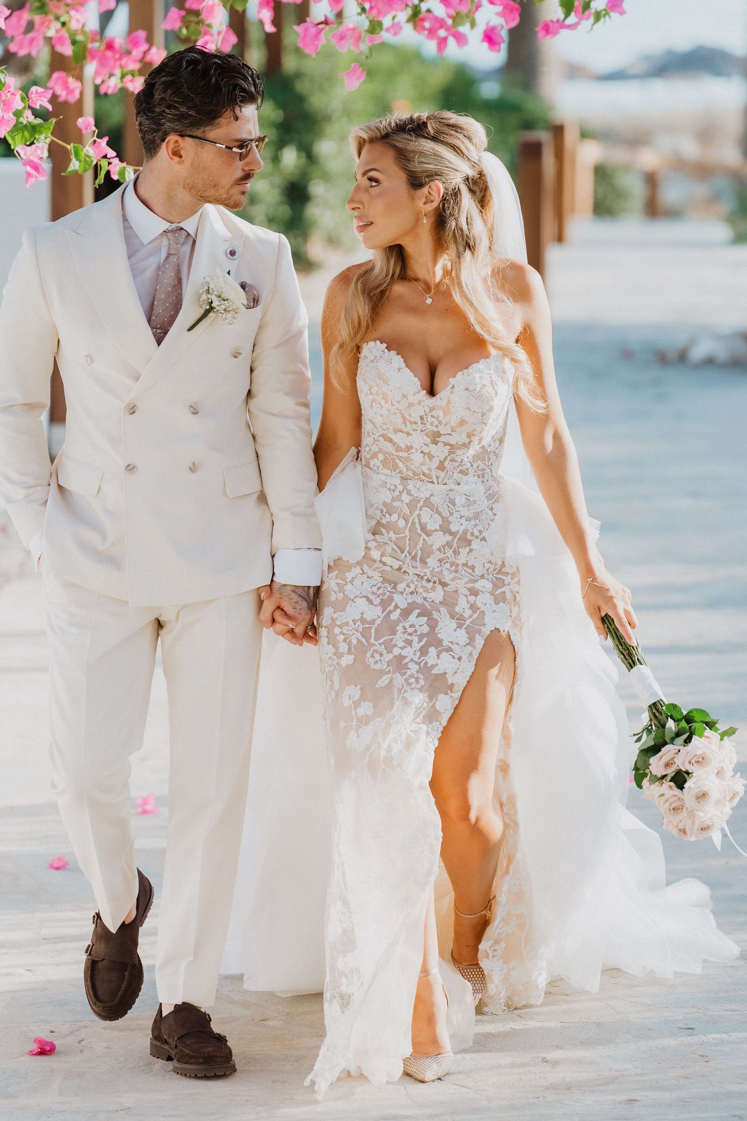 Newlywed couple walking together on a sunny day with pink flowers in the background.