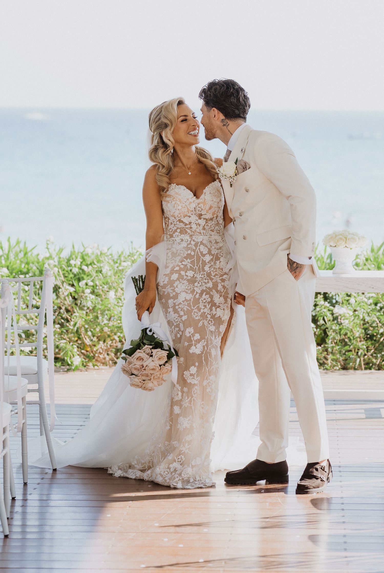 A couple shares a romantic moment at their beachside wedding, with the bride wearing a lace gown and groom in cream suit.
