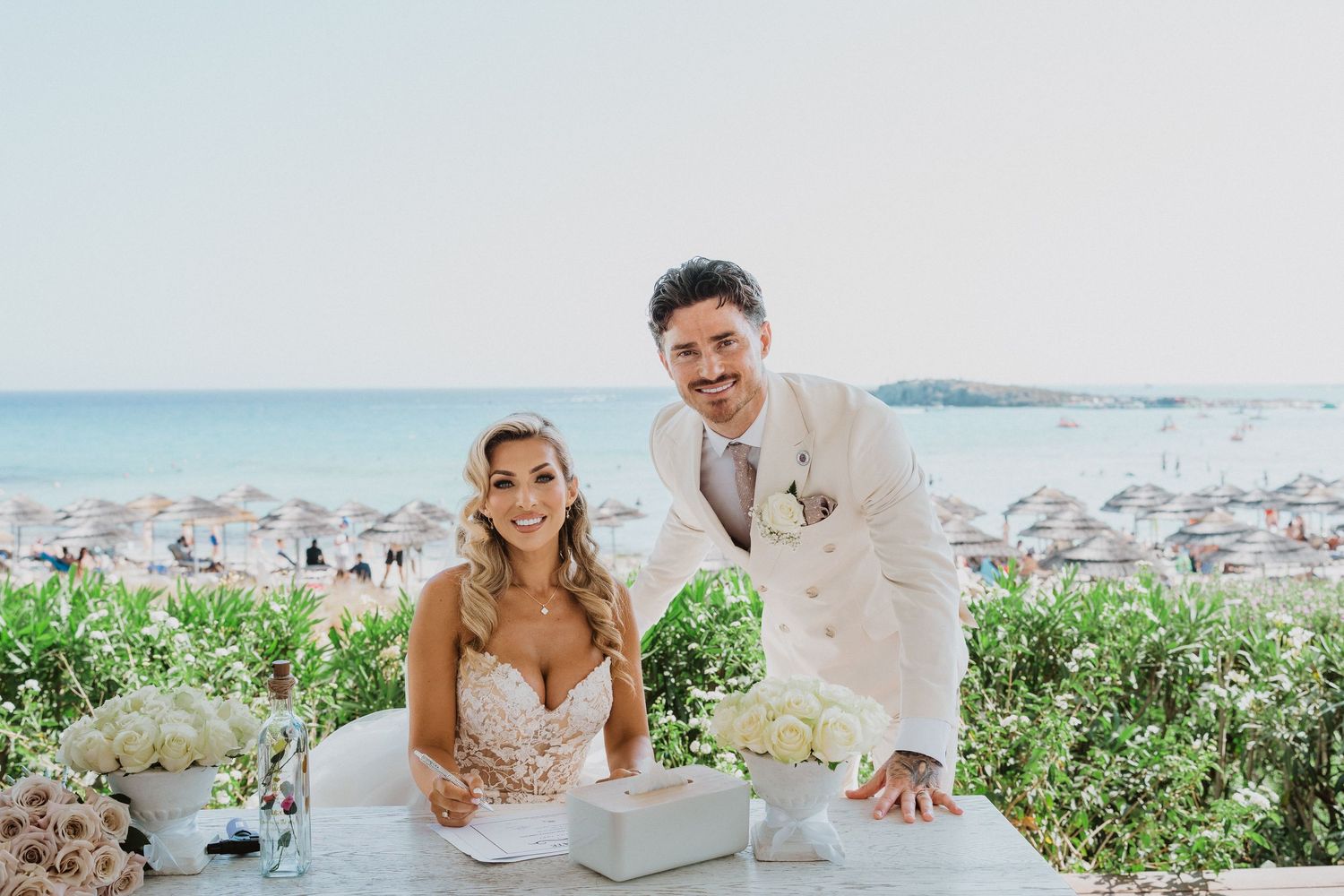 Couple poses together during romantic beach wedding photoshoot with ocean and greenery in background.