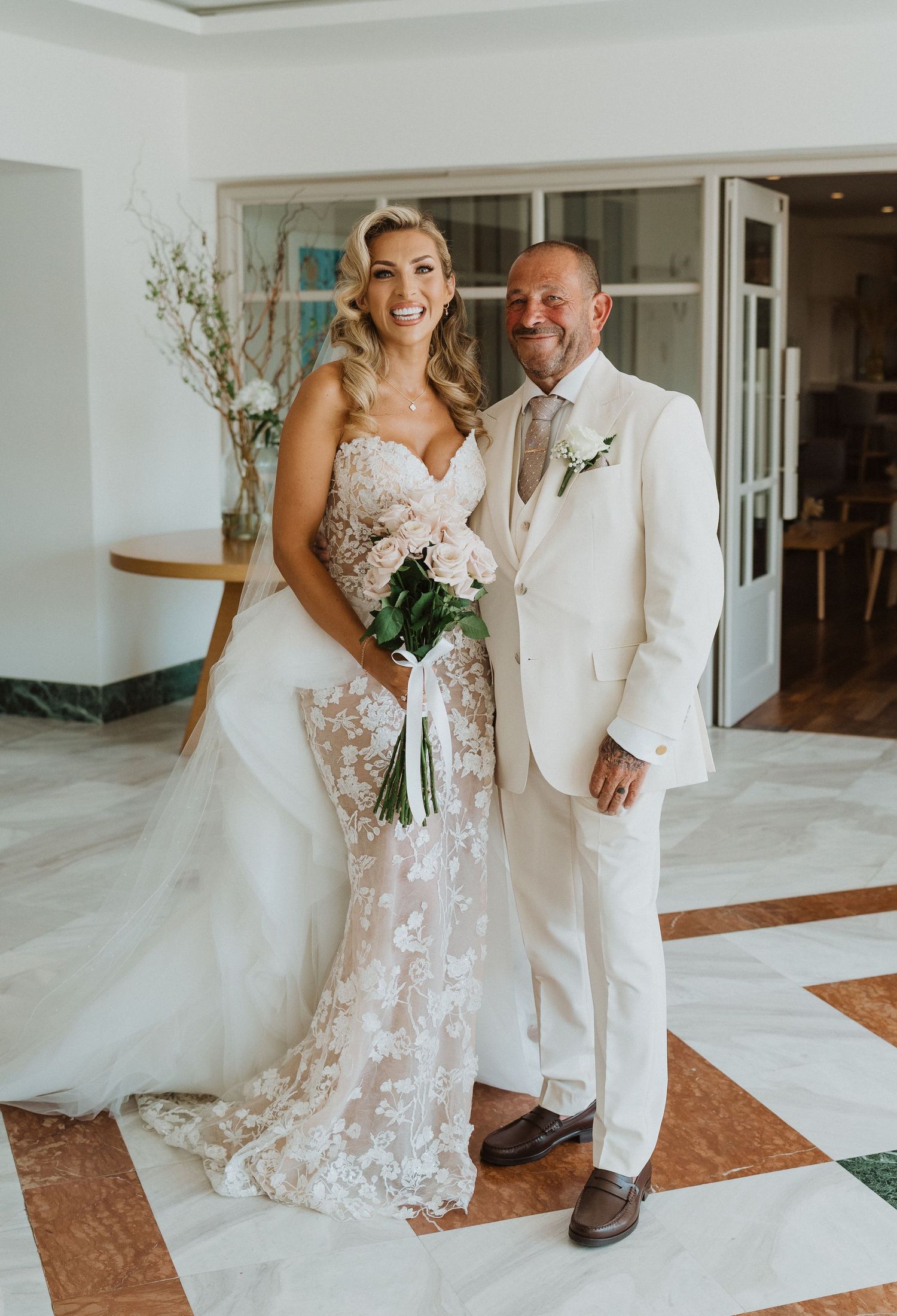 A couple in wedding attire shares a tender moment in an elegant indoor venue with white walls and marble floors.