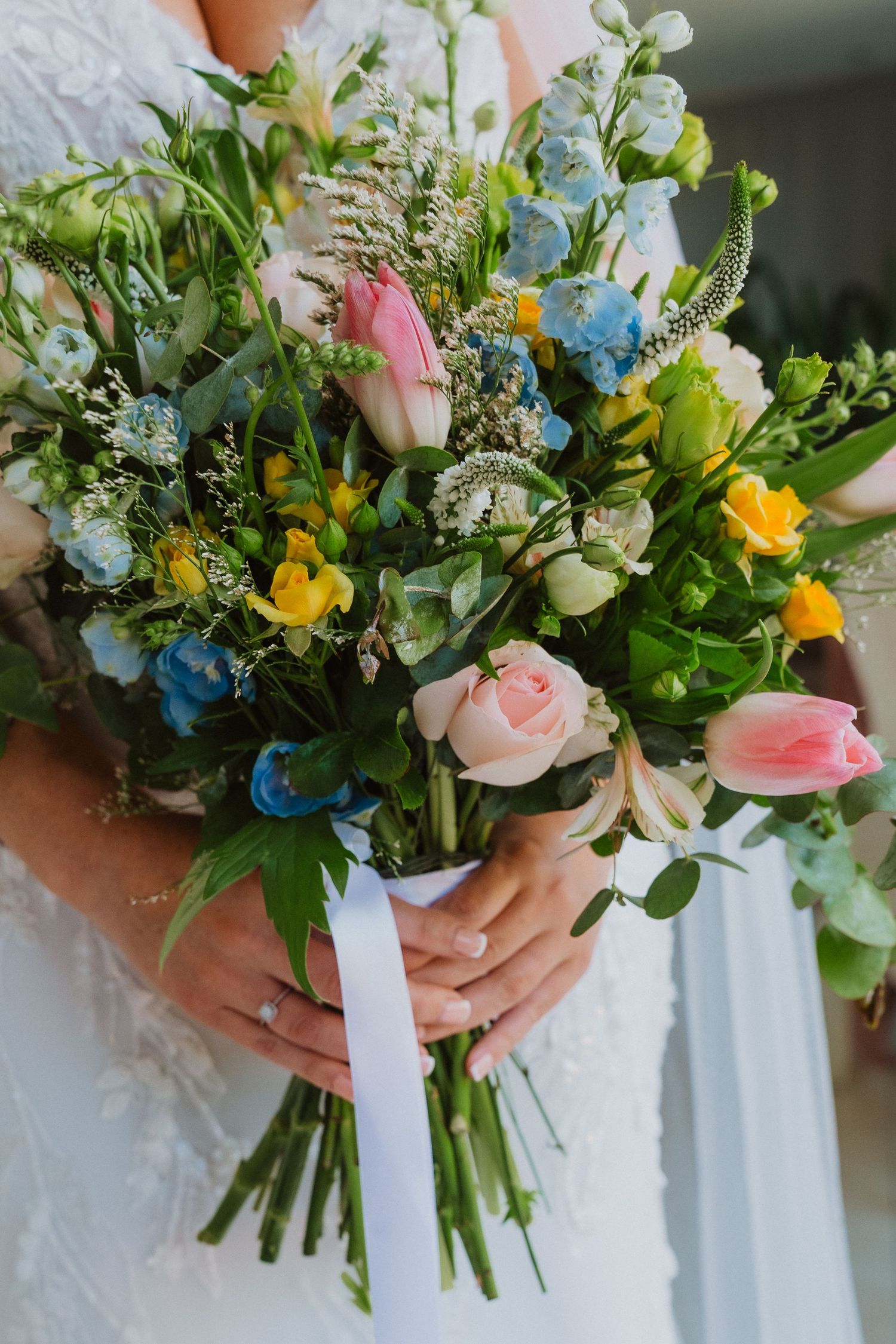 A series of wedding bouquets featuring pink roses, blue delphiniums, and greenery held against a white dress background.