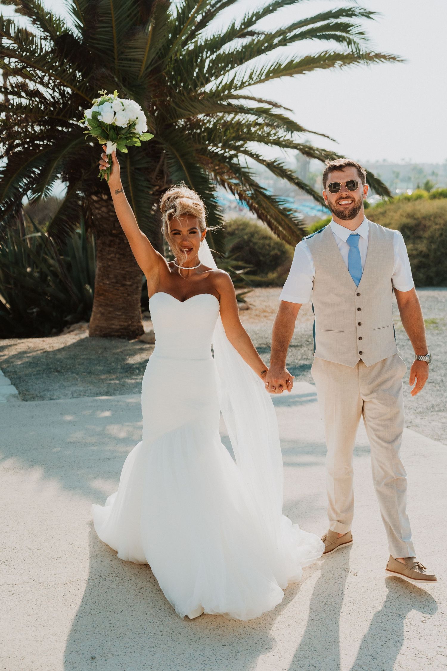 Joyful bride in white gown raises bouquet while groom in tan suit holds her hand near a palm tree.