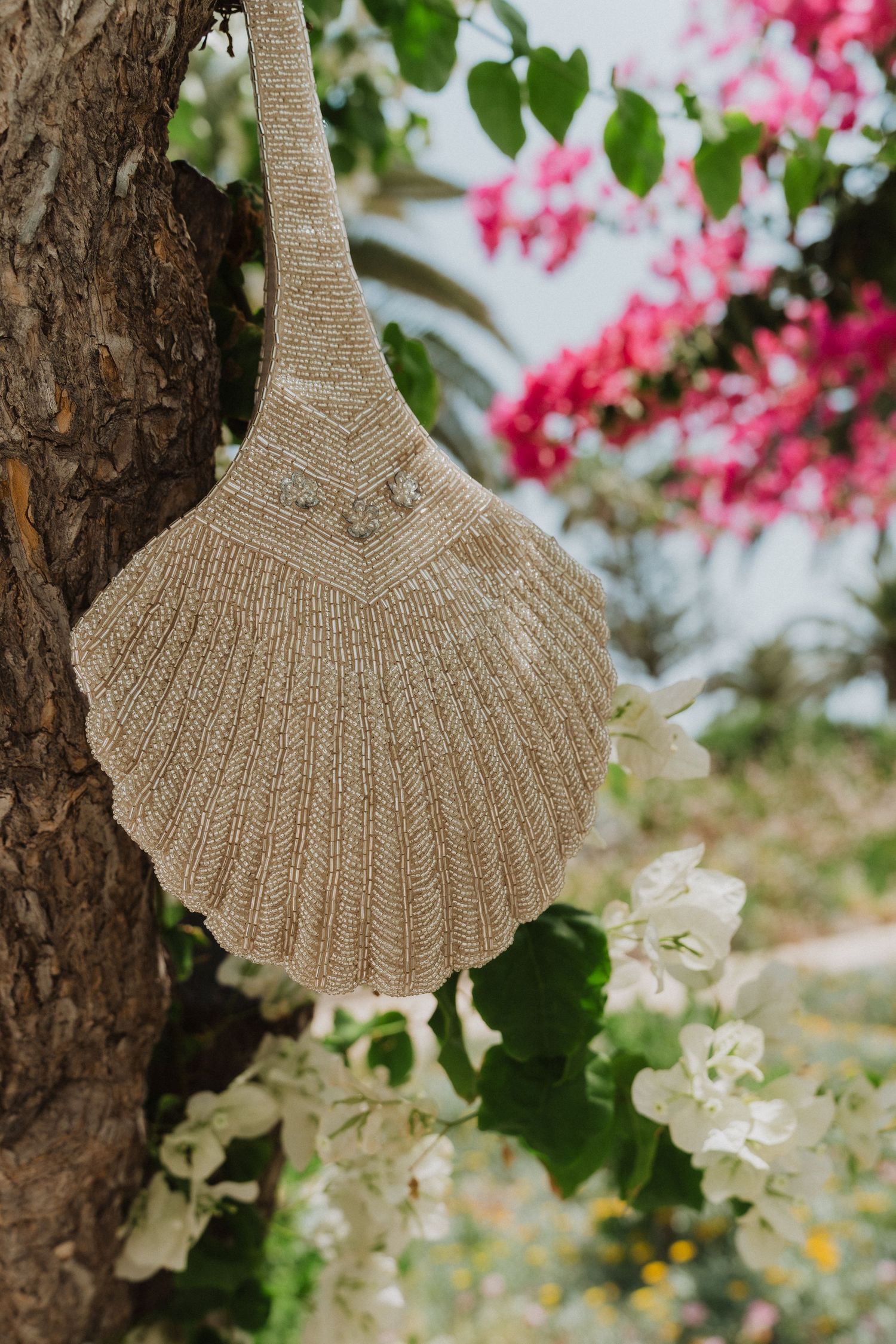 Woven seashell-shaped bridal bag hanging on a tree with pink flowers.
