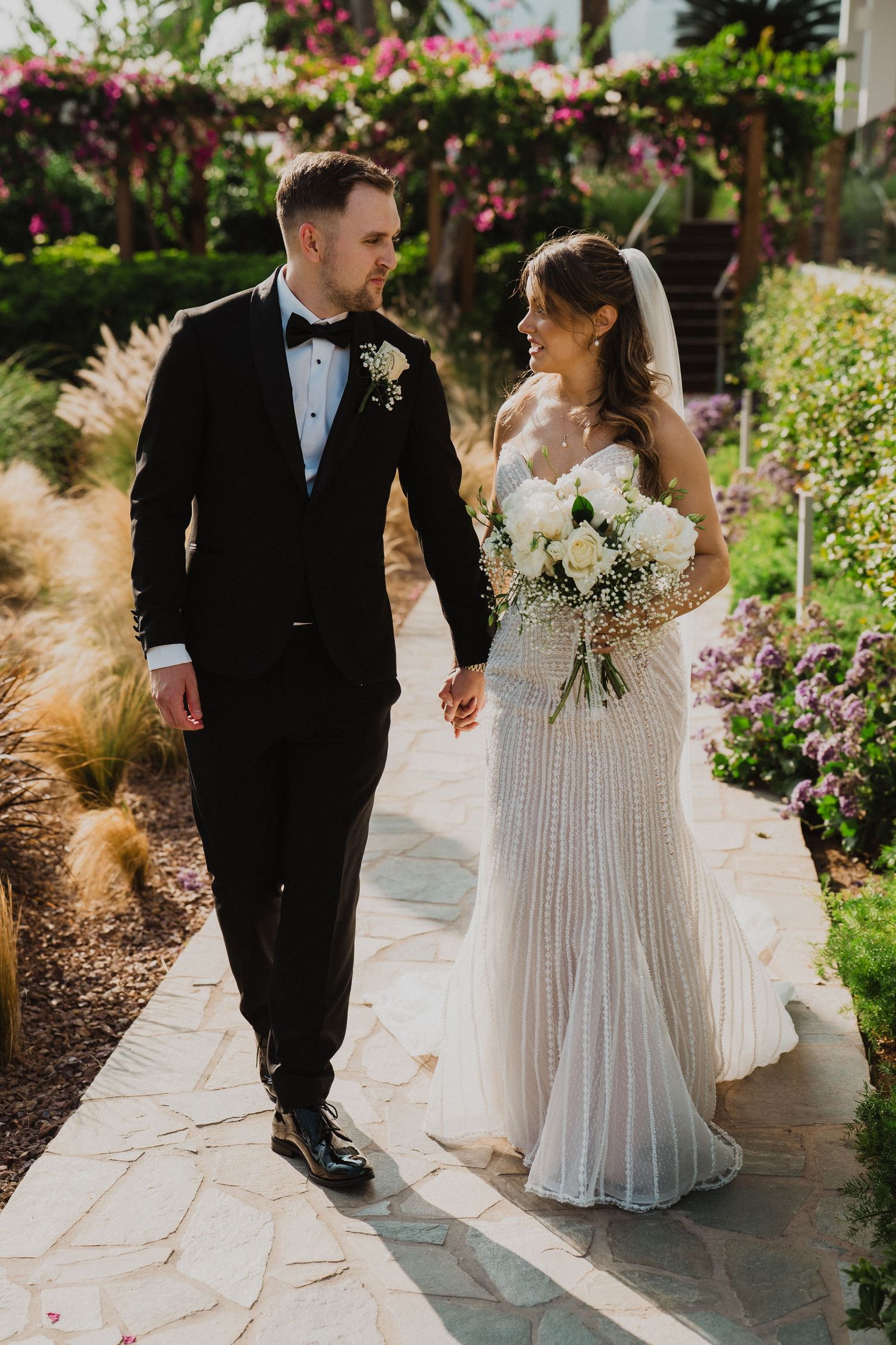 Bride and groom walk hand in hand through a lush garden path, she wearing a beaded gown holding white roses.