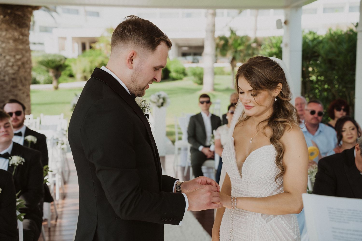 A groom in black suit places a wedding ring on his smiling bride in a beaded white gown during ceremony.