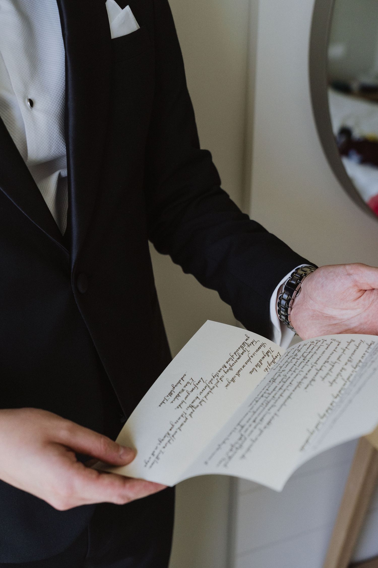 Groom in black tuxedo reading handwritten wedding vows before the ceremony.